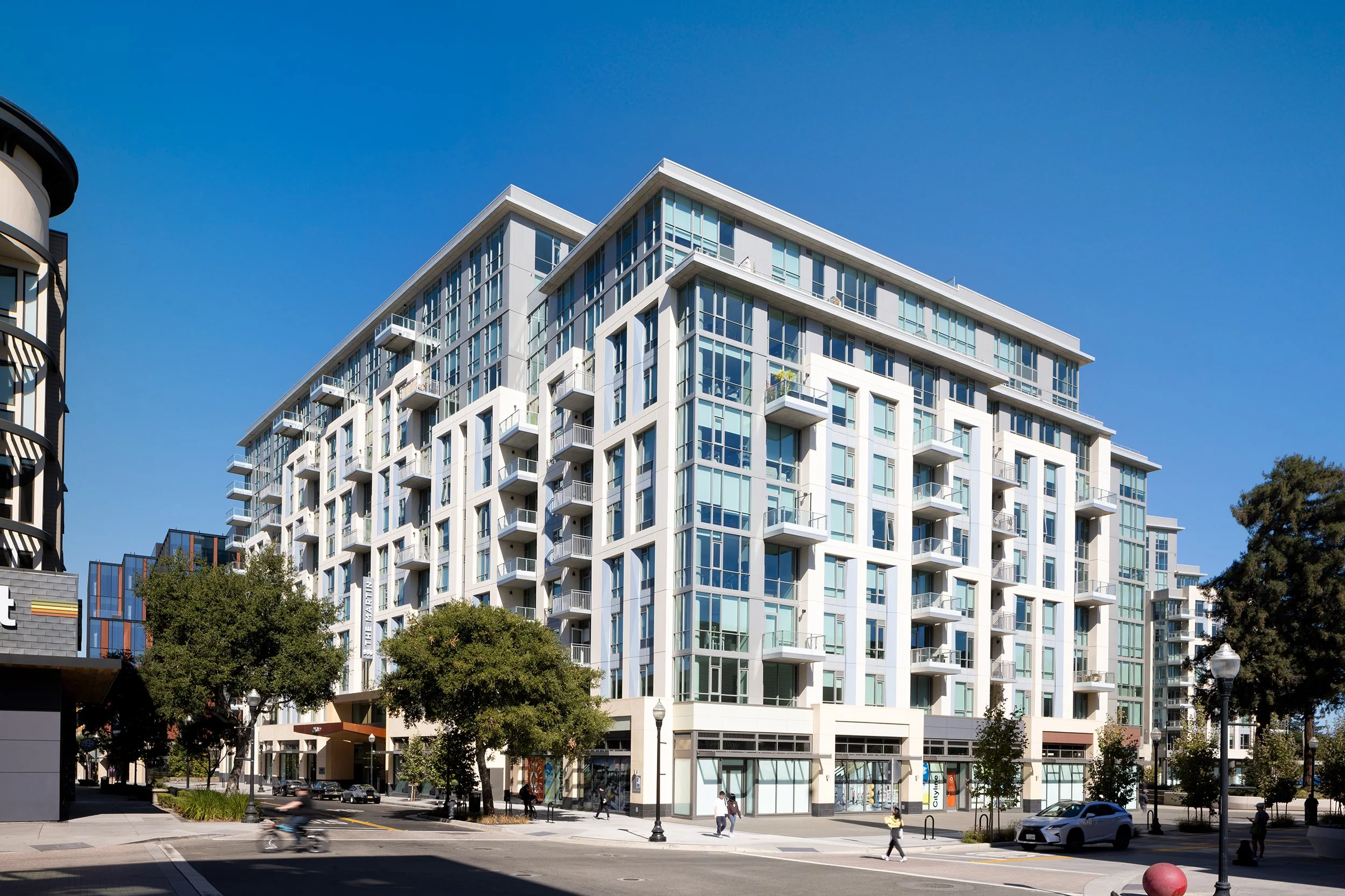 Modern multi-story residential building with large glass windows and balconies, situated on a city street with trees, pedestrians, and cars, under a clear blue sky.
