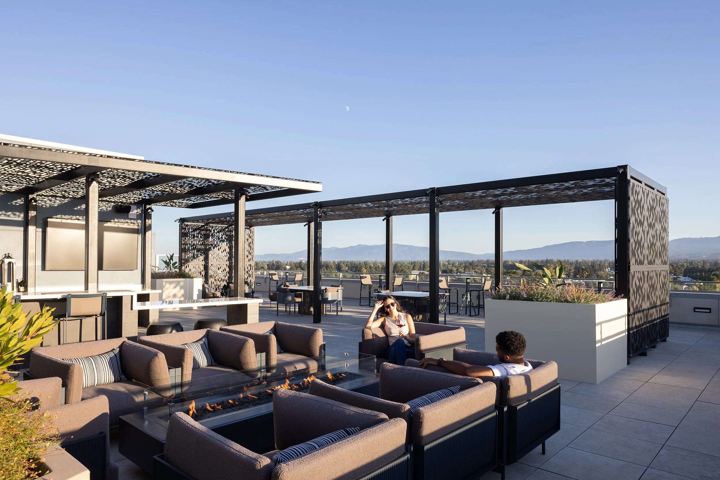 People relaxing on outdoor furniture on a rooftop patio with a view of distant mountains under a clear sky.