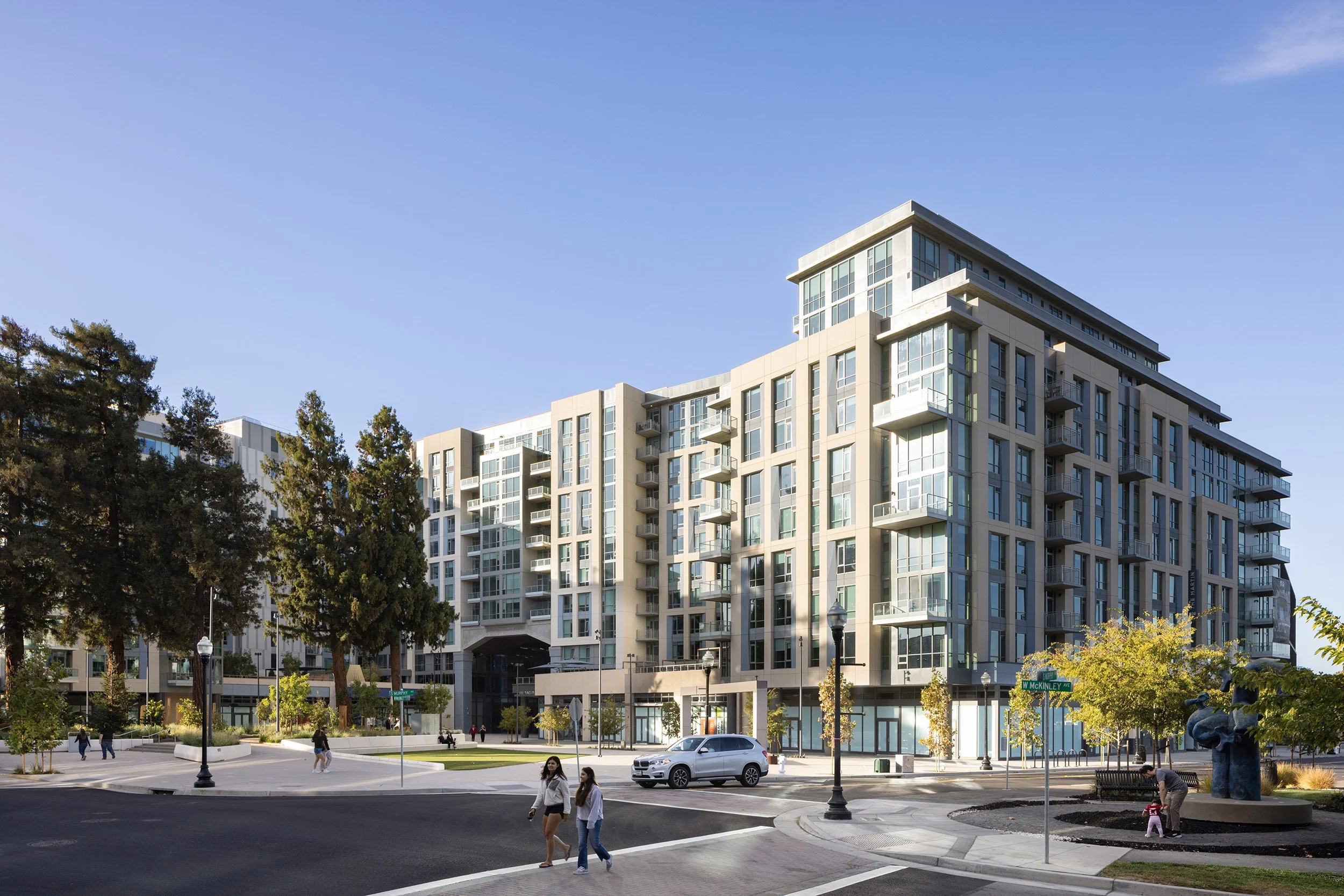Modern multi-story apartment building with glass balconies, surrounded by trees, at a city intersection with people walking, cars, and street lamps.