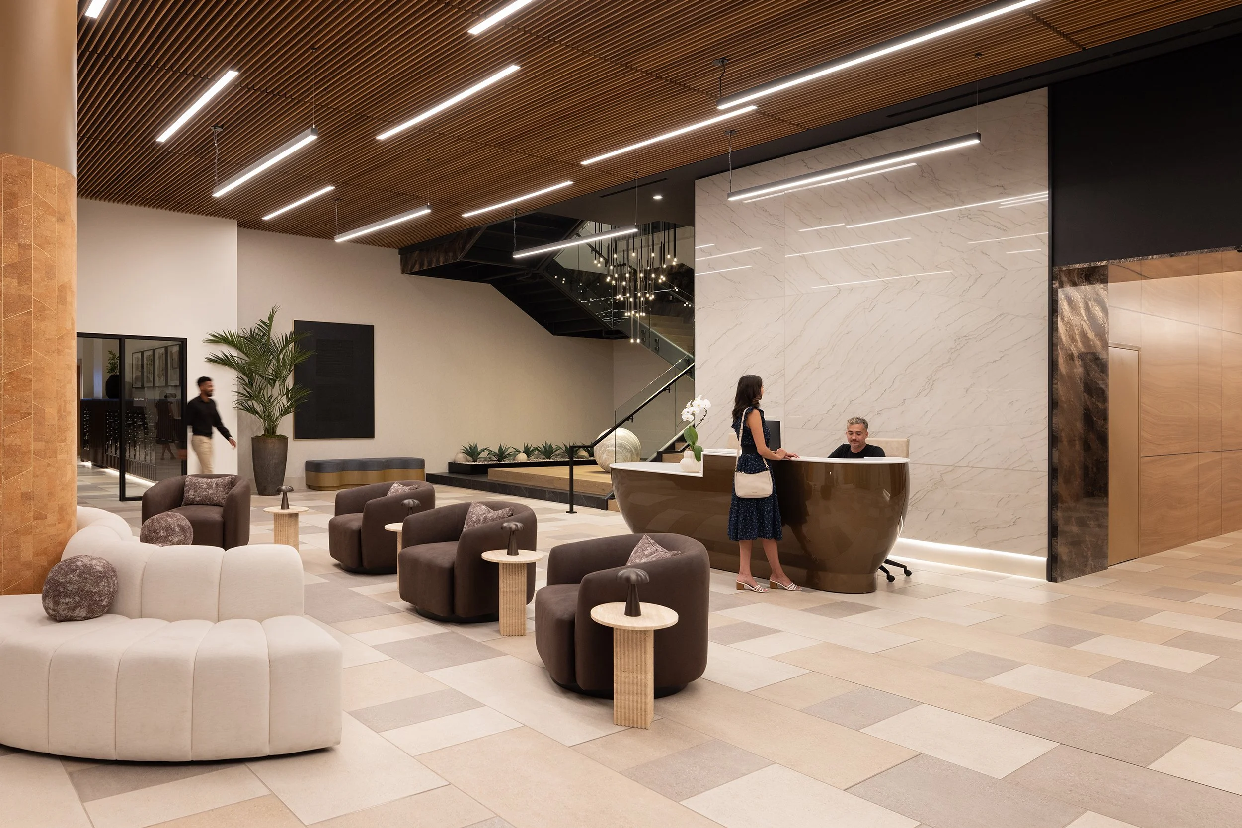 Modern hotel lobby with seating area featuring white and brown lounge chairs, small side tables, a reception desk with a woman checking in, a man behind the desk, decorative plants, and contemporary lighting fixtures.