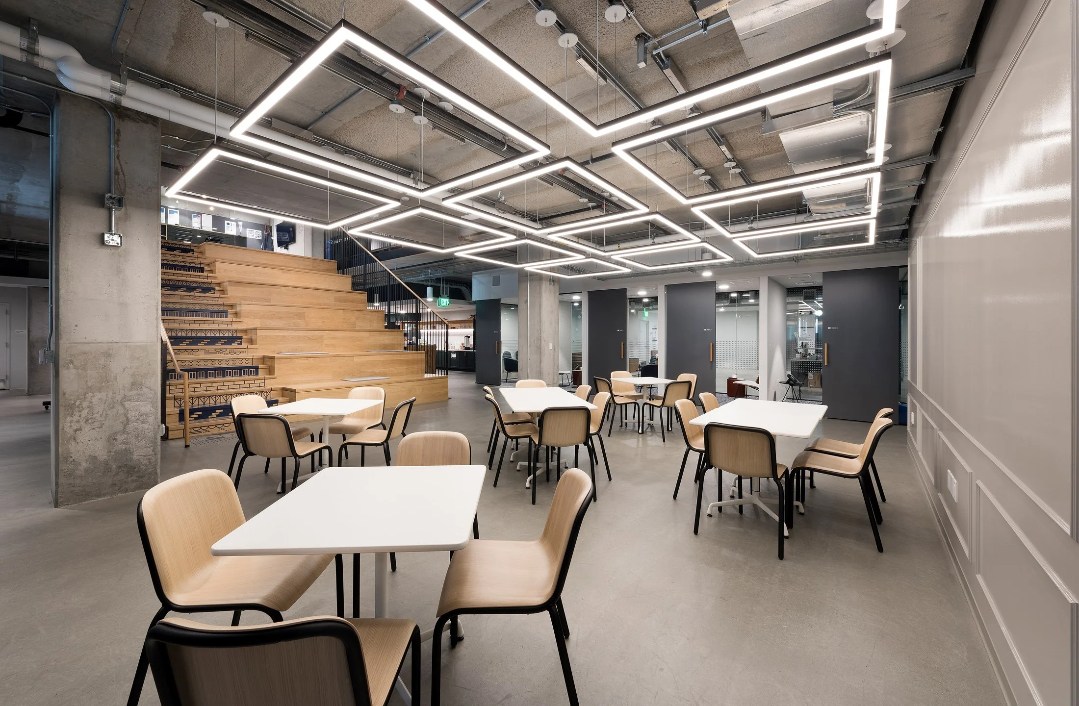 Modern office cafeteria with white tables, beige chairs, wooden stairs, and ceiling lights.