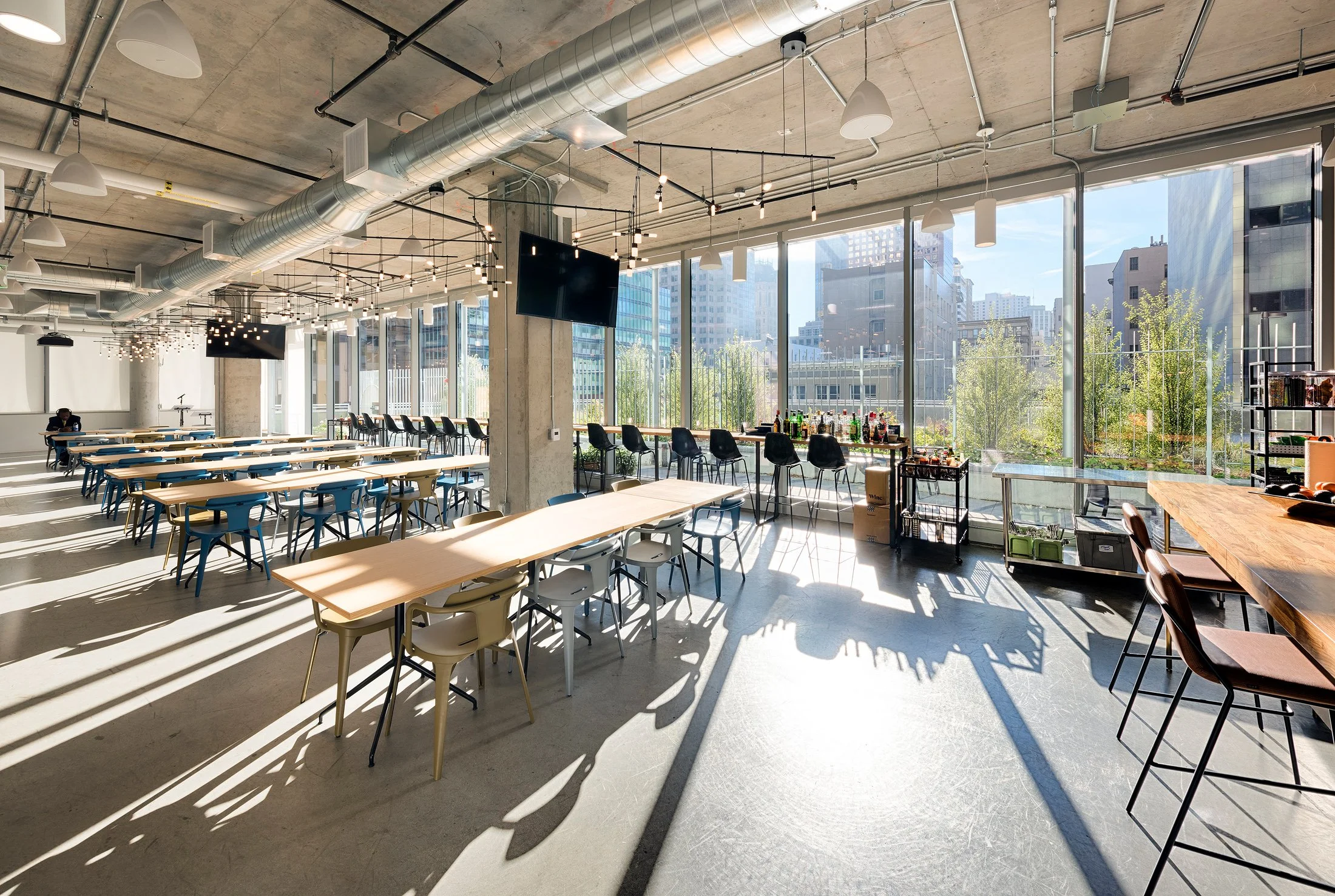 A spacious modern conference room with large windows, sunlight streaming in, and a cityscape view outside. The room has multiple tables and chairs, some arranged for meetings or events, with a bar area along the window.