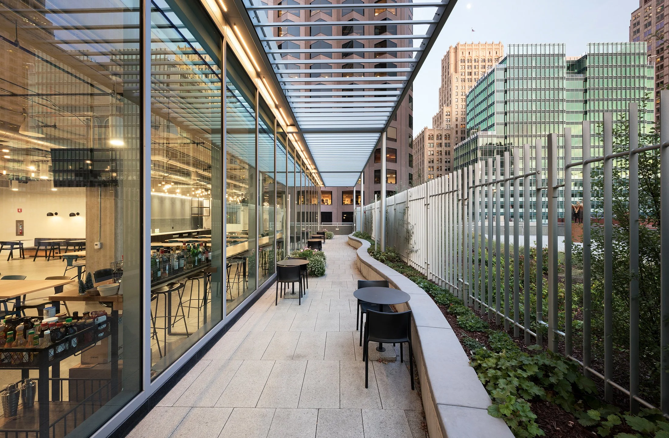 An outdoor balcony area of a restaurant or cafe with small round black tables and chairs, glass walls revealing the interior, and high-rise buildings in the background, during daytime.