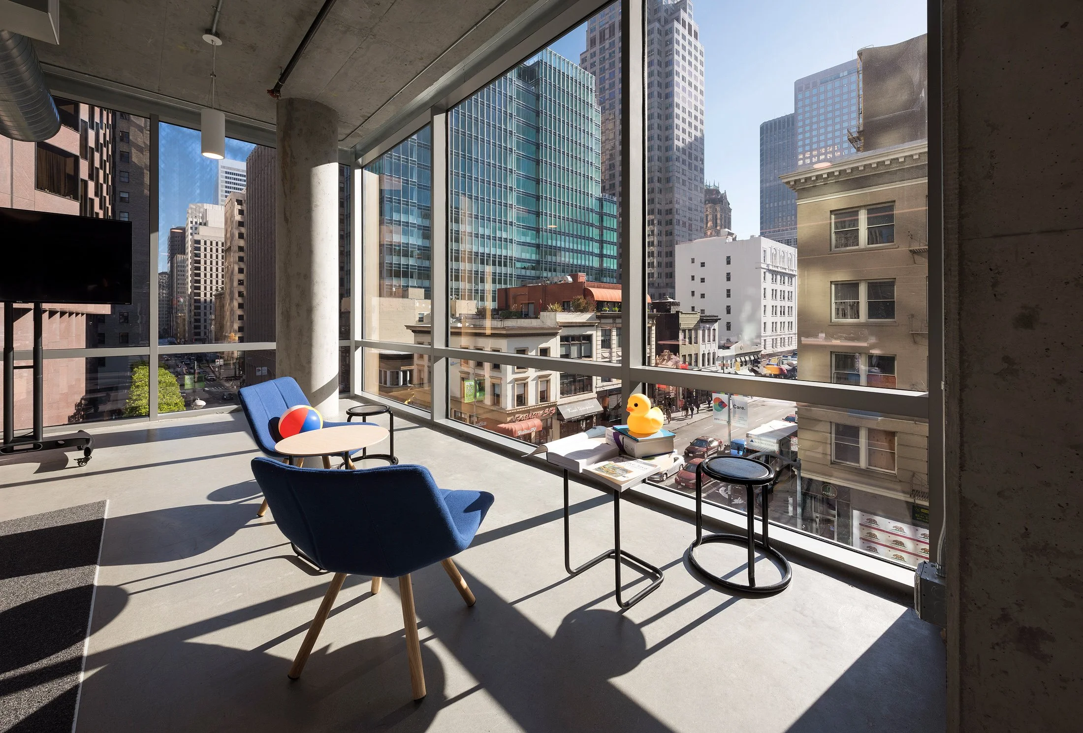 Modern office space with large windows overlooking a city street, featuring two blue chairs, a small round table, and decorative items including a beach ball, a rubber duck, and books.