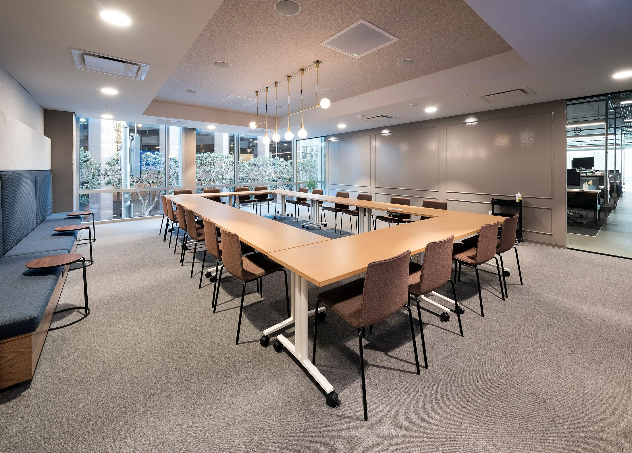 Modern conference room with a U-shaped arrangement of wooden tables, brown chairs, a blue cushioned bench with small tables, large glass windows with views of trees outside, and office space visible through a glass wall on the right.