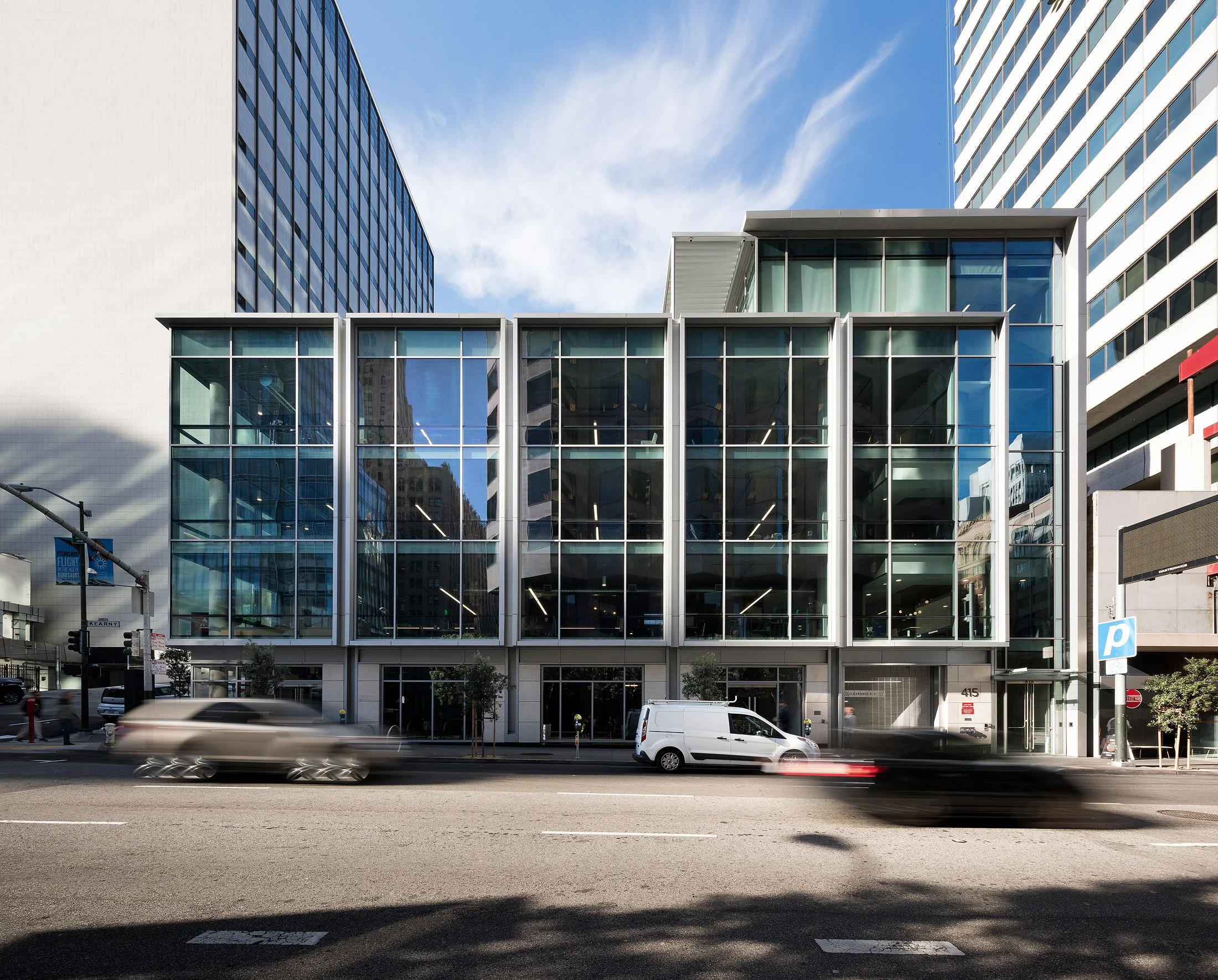 Modern glass-fronted building on city street with blurred cars passing by and tall office buildings in background.