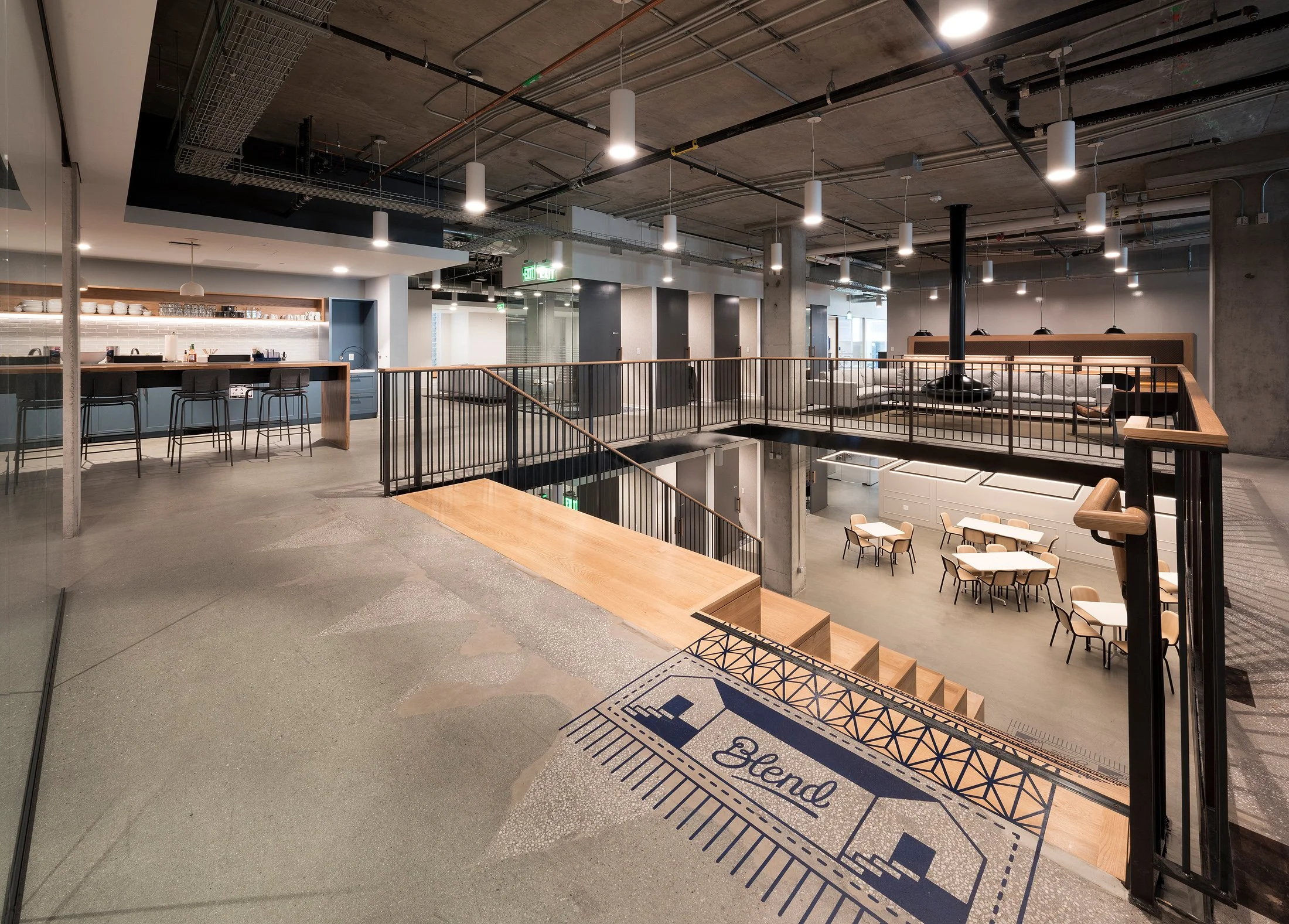 Modern, industrial-style interior of an office or co-working space with open layout, concrete walls, hanging light fixtures, a bar area with stools on the upper level, and casual seating areas on both levels.
