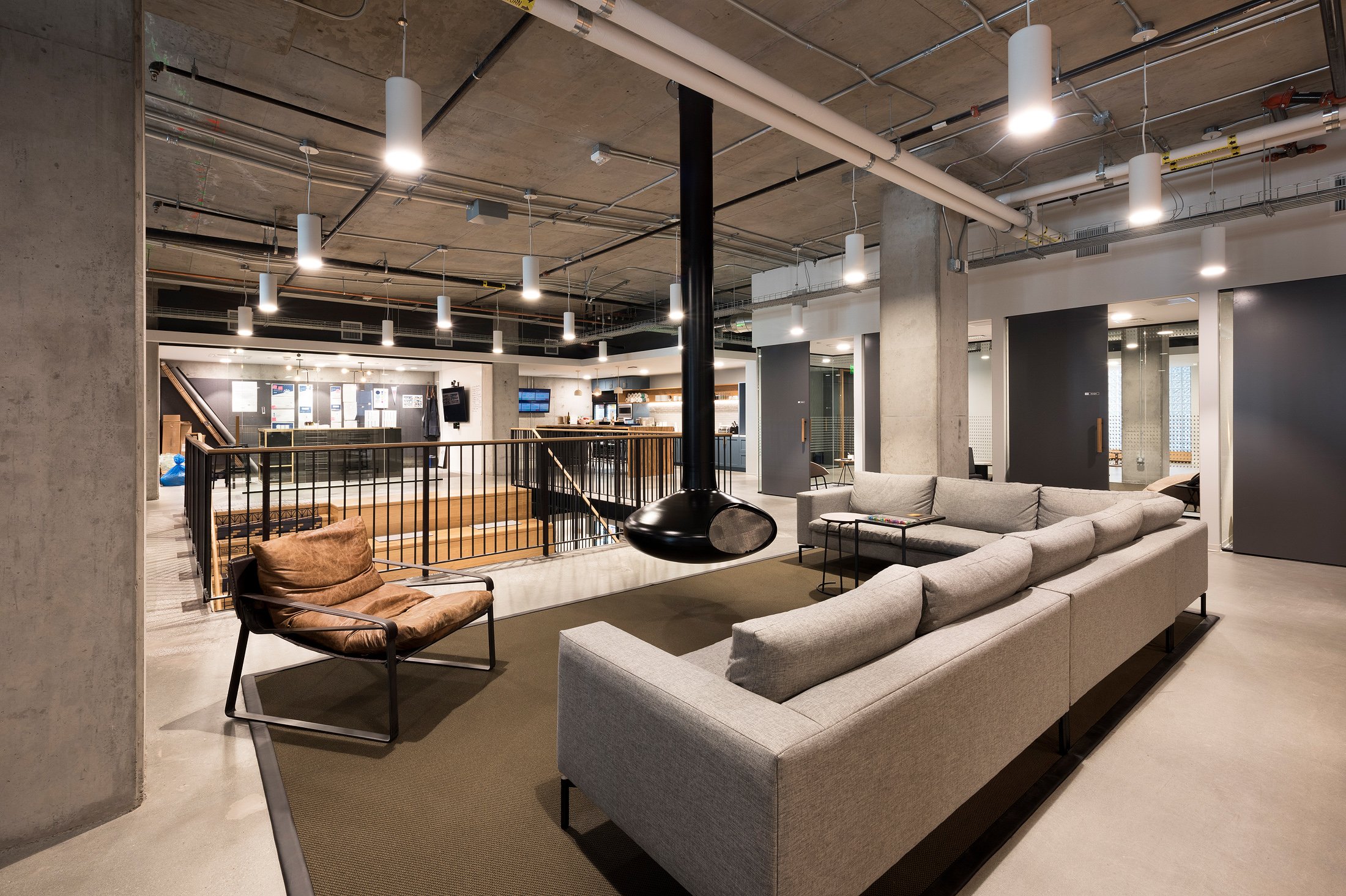 Modern industrial lounge area with gray sectional sofa, leather armchair, suspended black fireplace, and open view of a staircase and reception desk in the background.