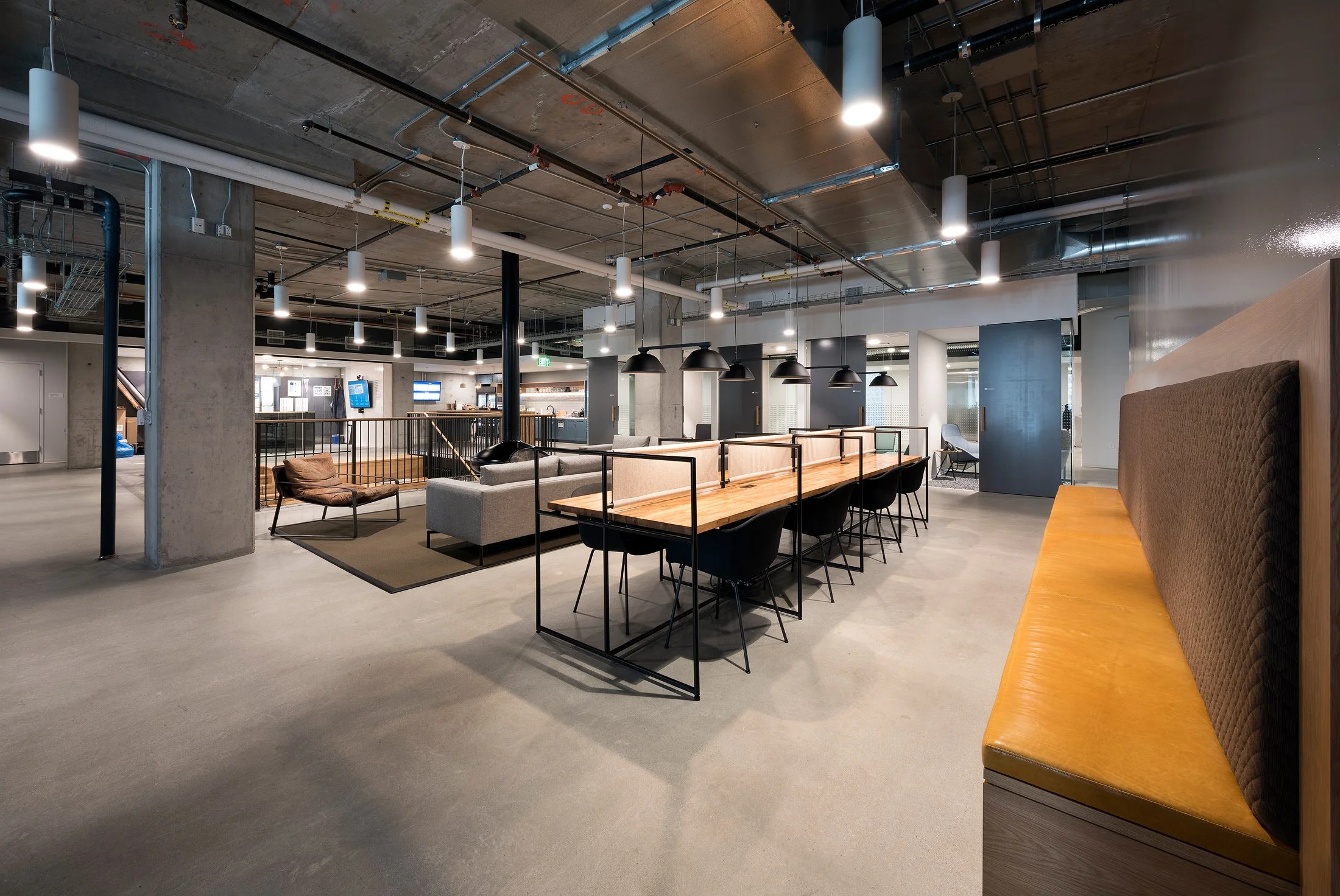 Modern, open-plan office lounge area with seating, a long communal table with black chairs, and a mix of cozy chairs and sofas. Industrial ceiling with exposed pipes and ducts, and minimalistic lighting.