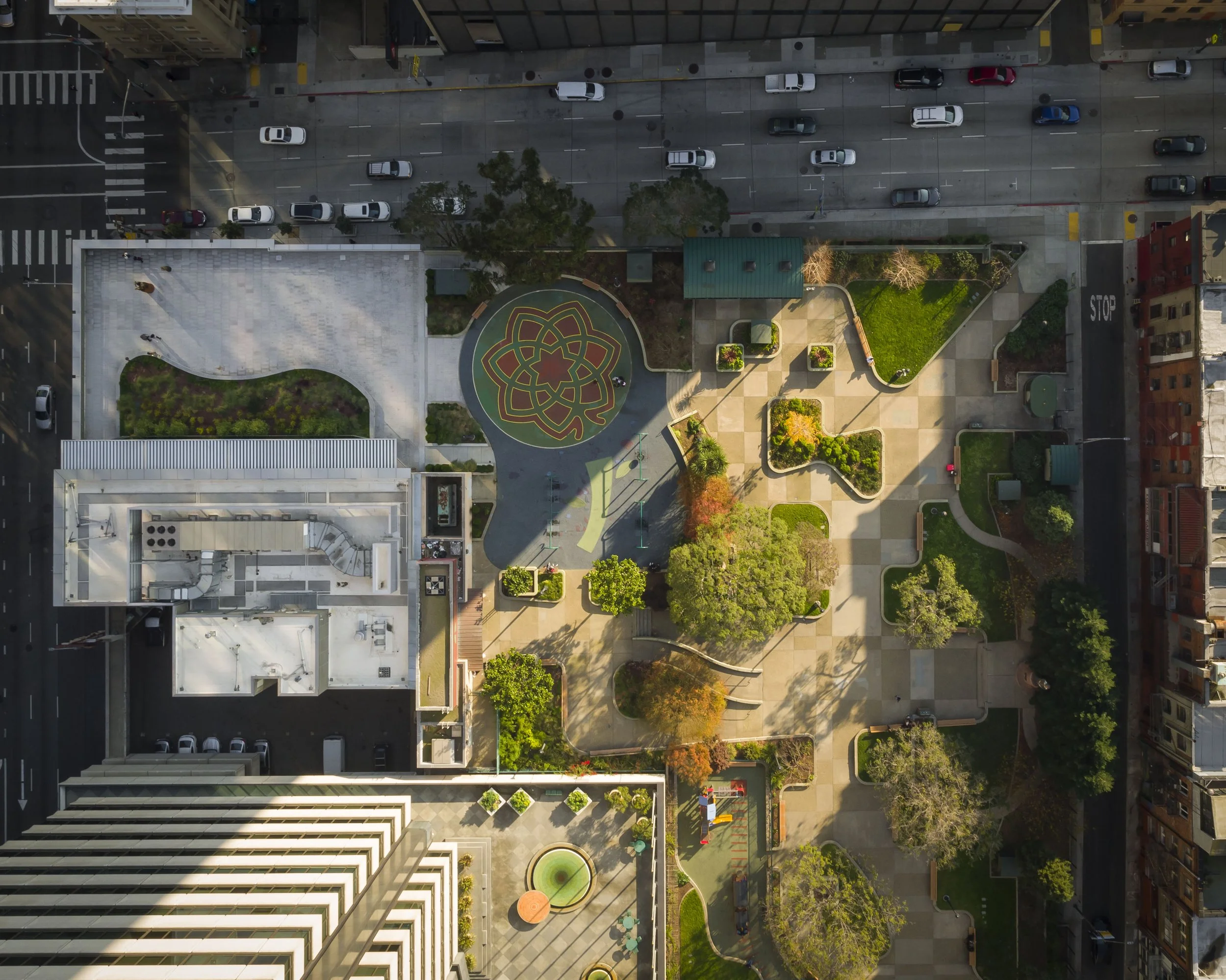 An aerial view of a city park with pathways, trees, and landscaping, surrounded by buildings and streets with parked cars.