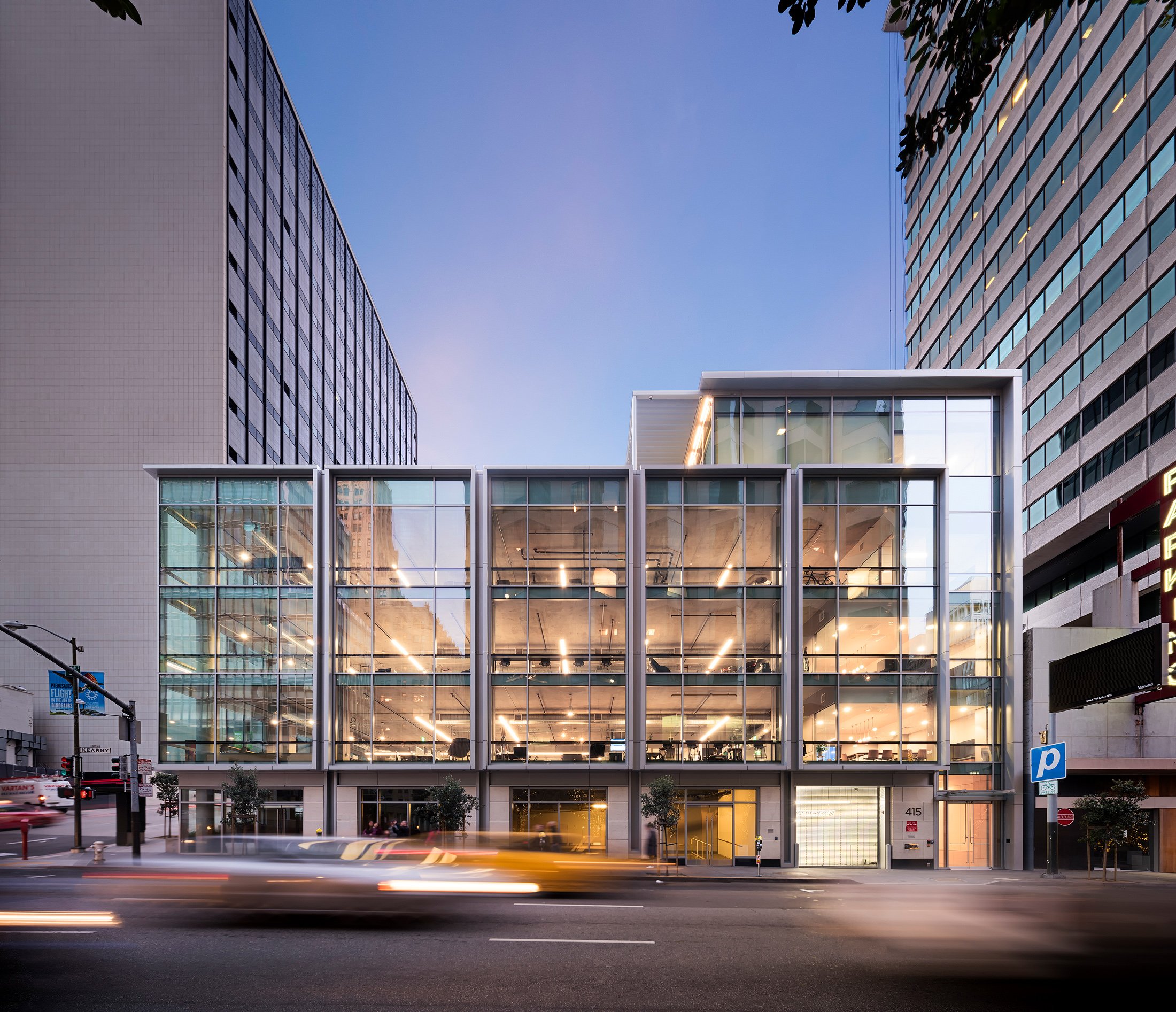 Modern multi-story glass building on city street during dusk, with streaks of light from moving cars in foreground, surrounded by tall skyscrapers.