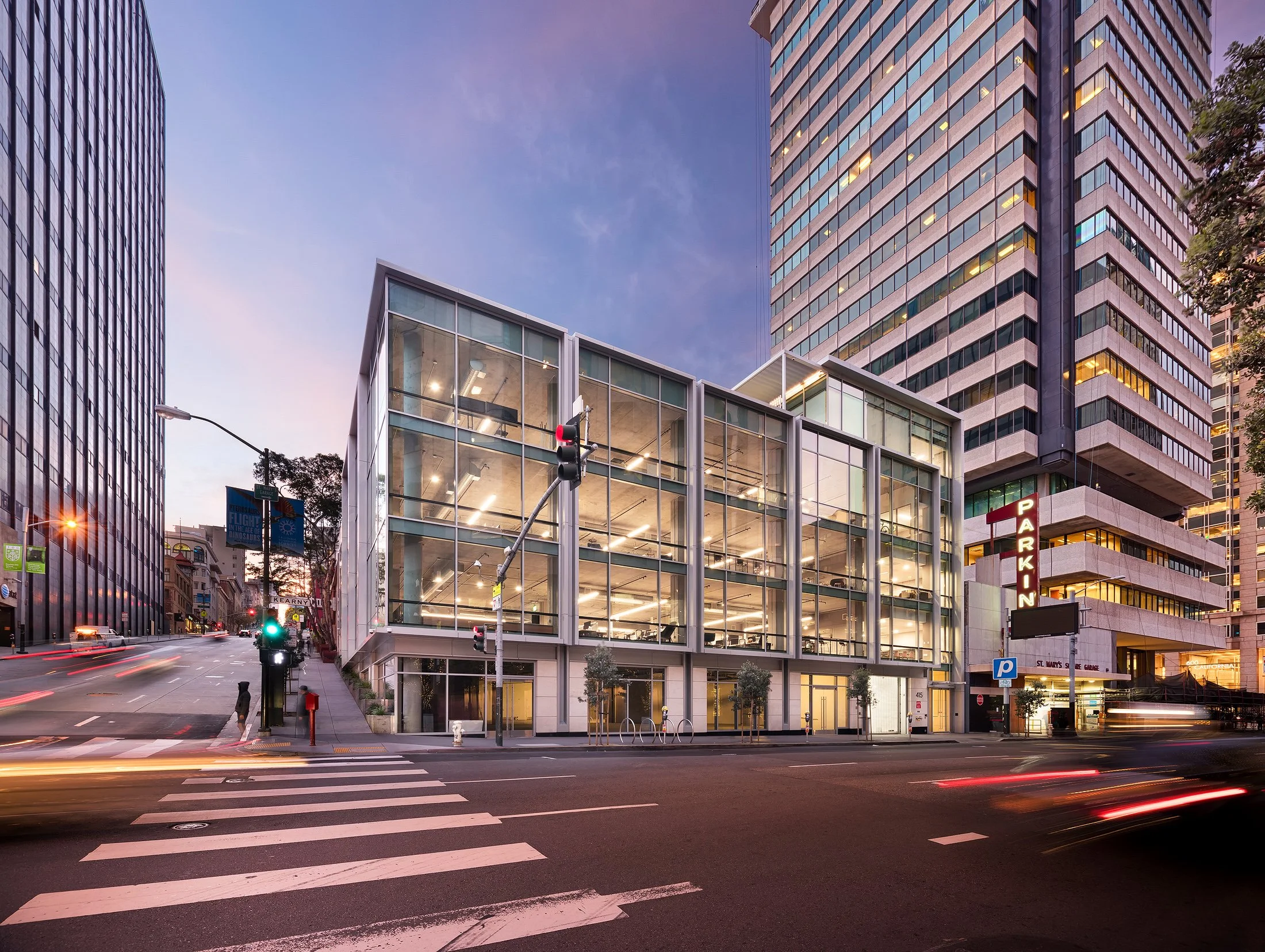 A modern glass building on a city street at dusk, with surrounding tall office buildings, traffic lights, and streaks of car lights from passing vehicles.