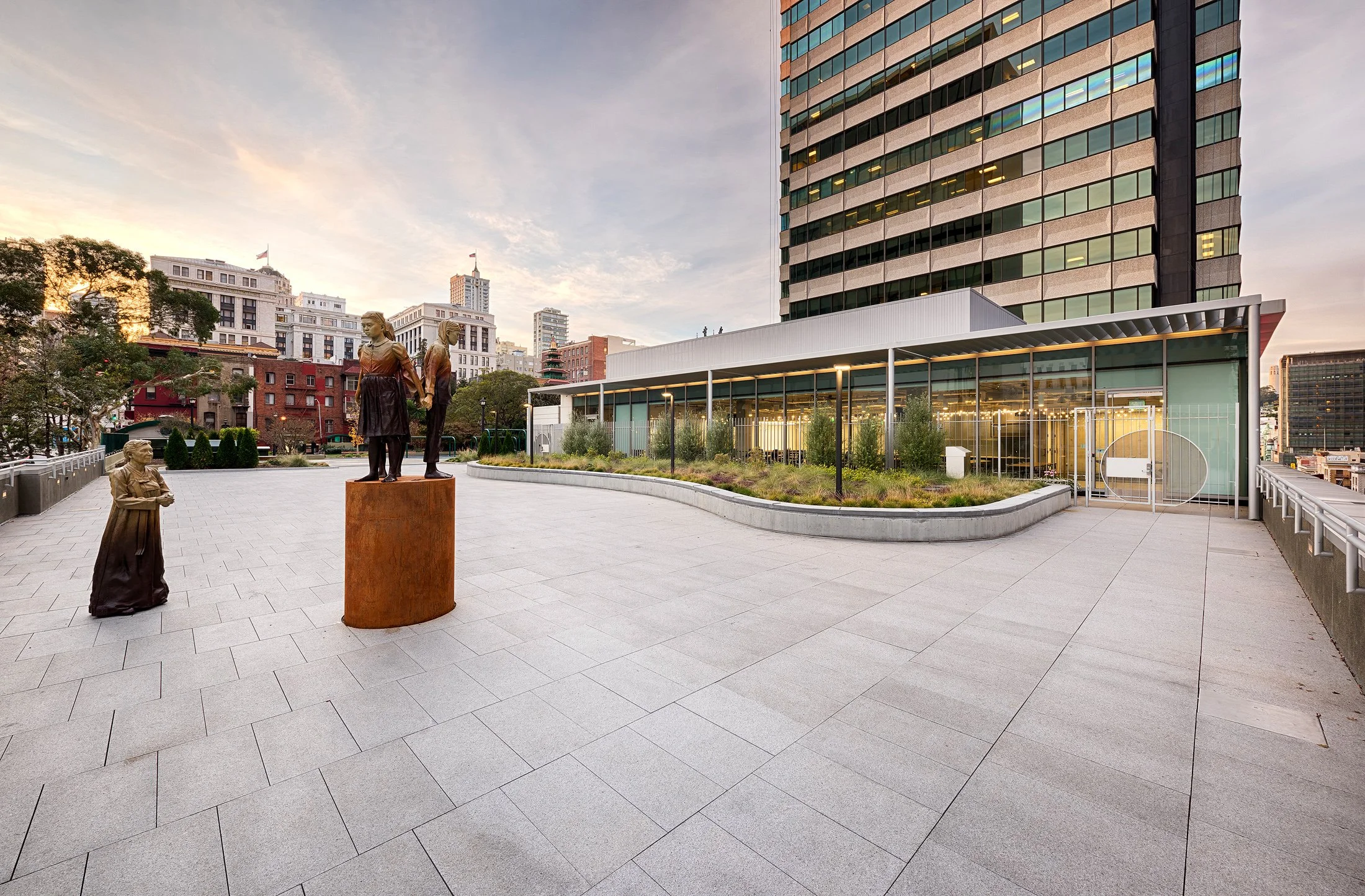Empty rooftop terrace with bronze statues of children, a modern building with glass walls, and city high-rise buildings in the background during sunset.