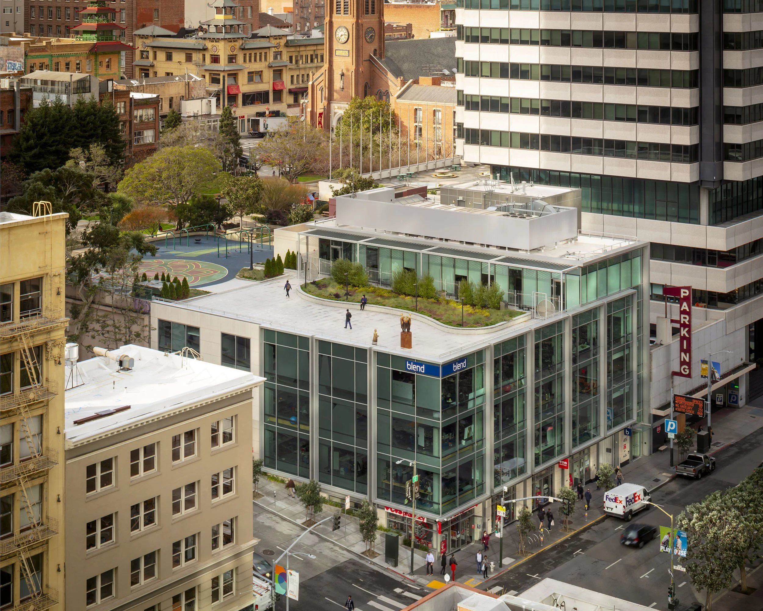 A cityscape view of a modern glass building with a rooftop garden, nearby older buildings, and a park with trees and a playground. Pedestrians are walking on the streets and a FedEx delivery truck is parked outside the building.