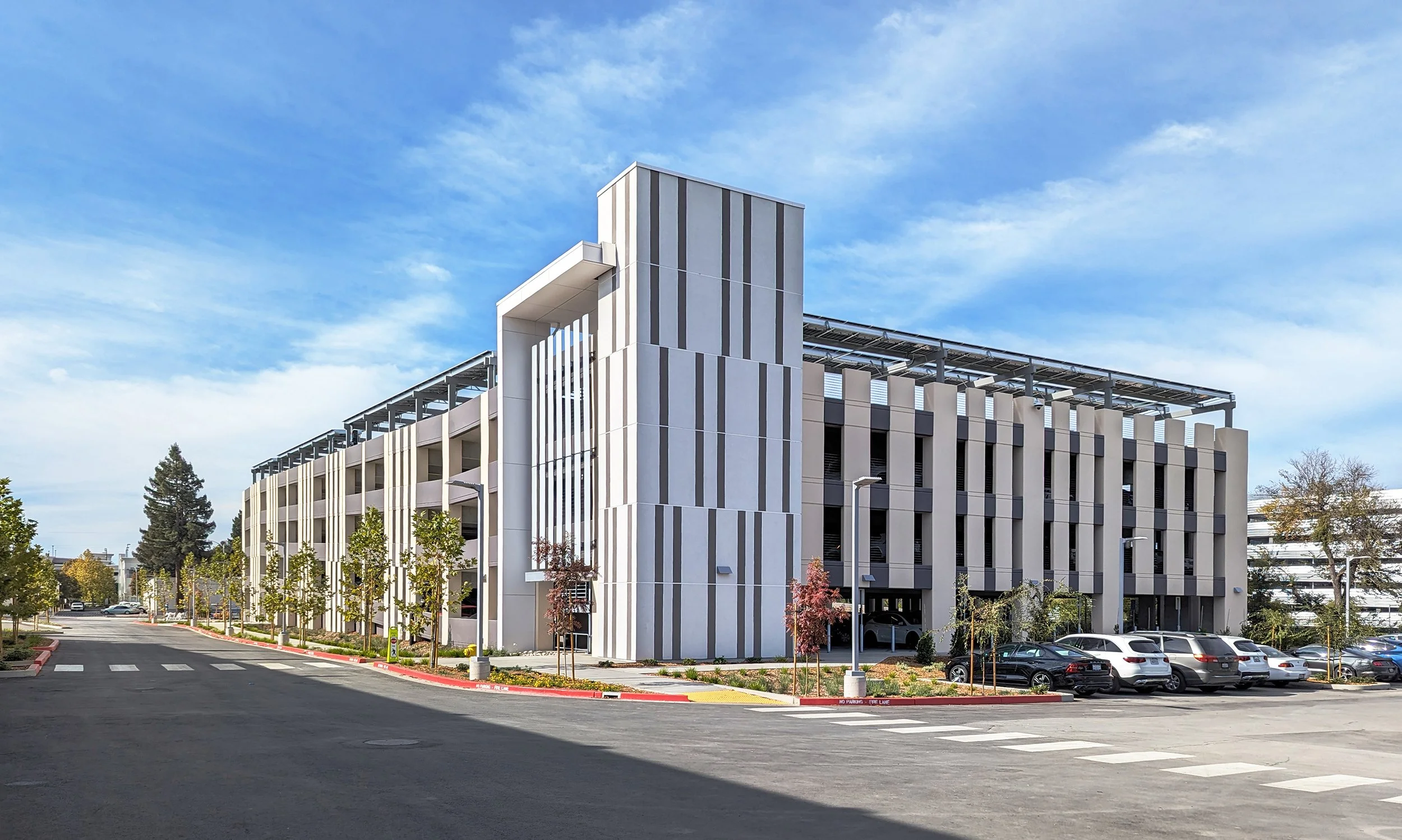 Modern multi-story parking garage with a geometric design, surrounded by parked cars, trees, and a clear blue sky.