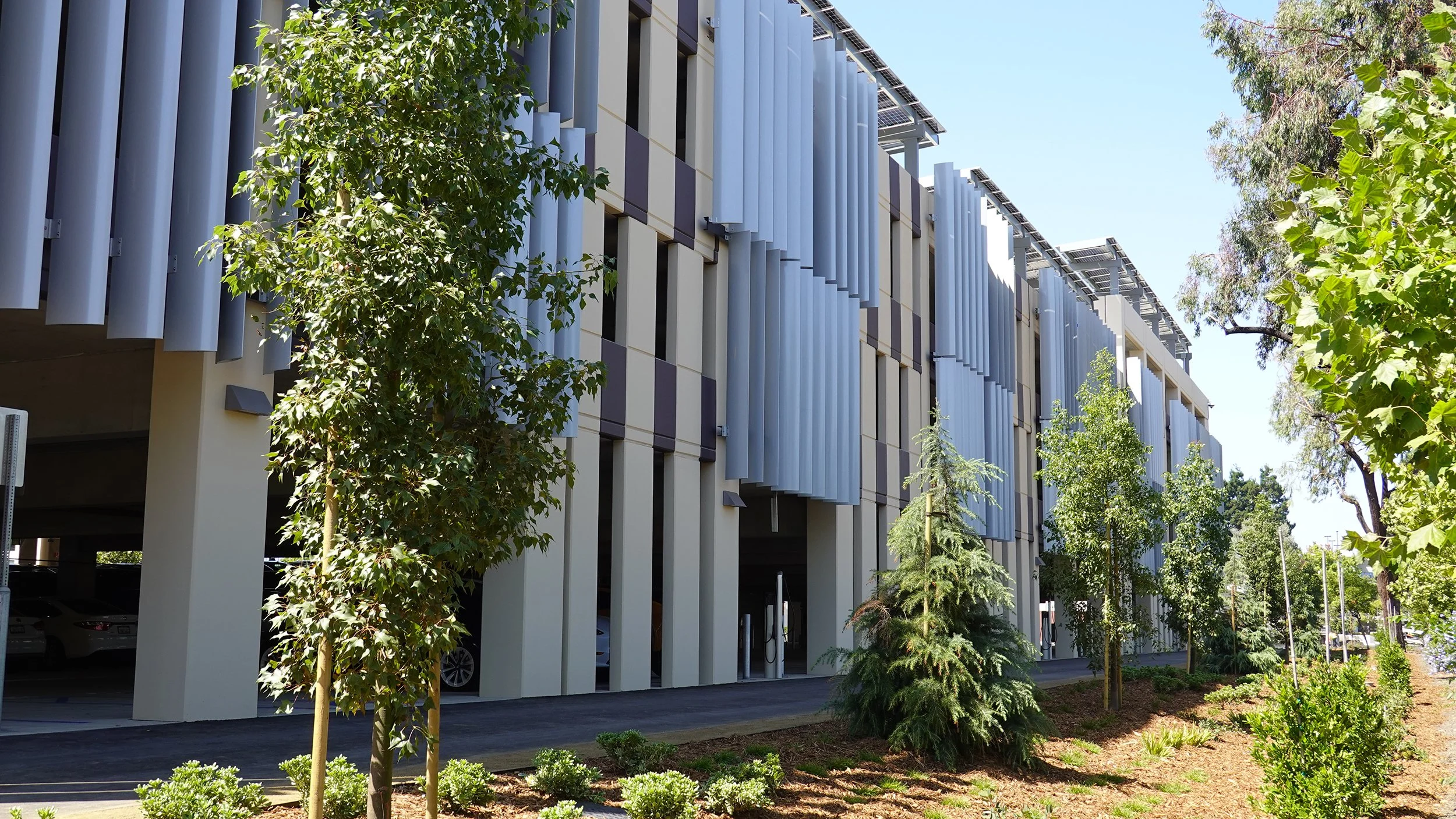 Modern building with vertical metal slats and surrounding greenery, including trees and shrubs, under a clear blue sky.
