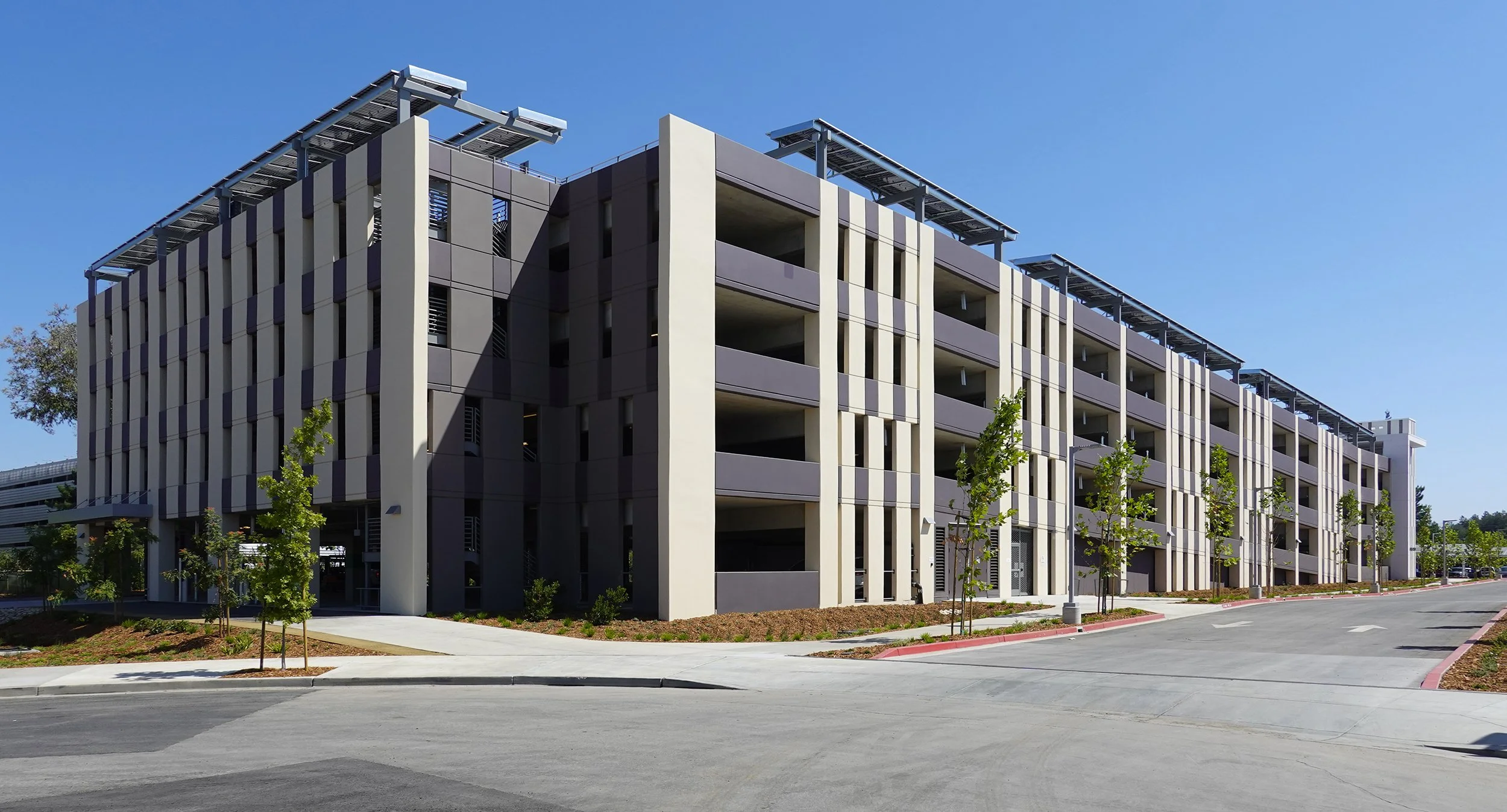 Modern multi-story parking garage with solar panels on the roof, surrounded by small trees and clear blue sky.