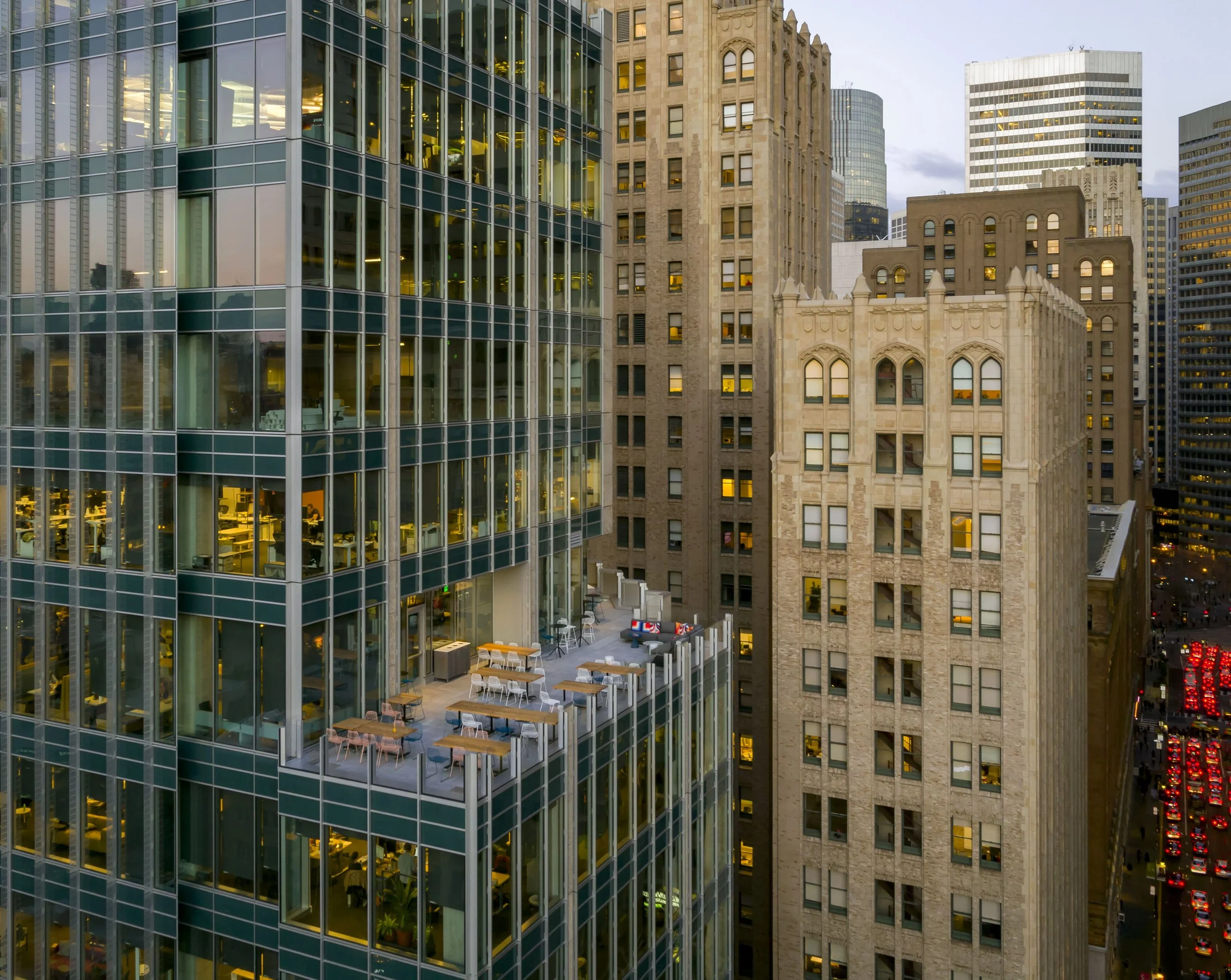 Cityscape featuring modern glass office building with outdoor terrace and older stone buildings, with traffic on the street below at dusk.