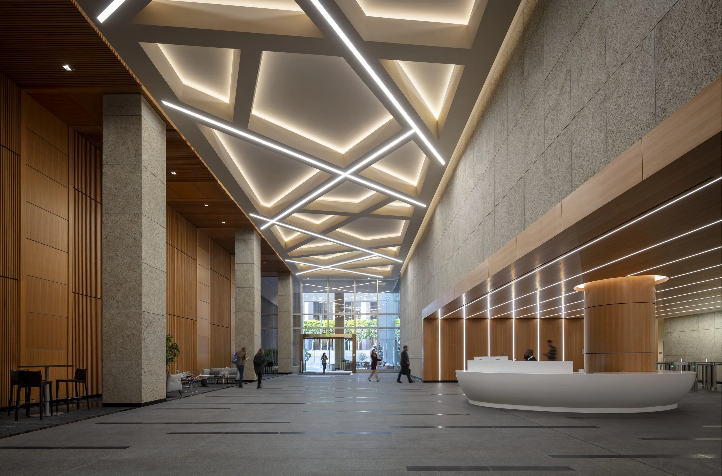 Modern hotel lobby with geometric ceiling lighting, wooden wall panels, and a reception desk with a curved design, several people walking or sitting, and glass doors at the back letting in natural light.