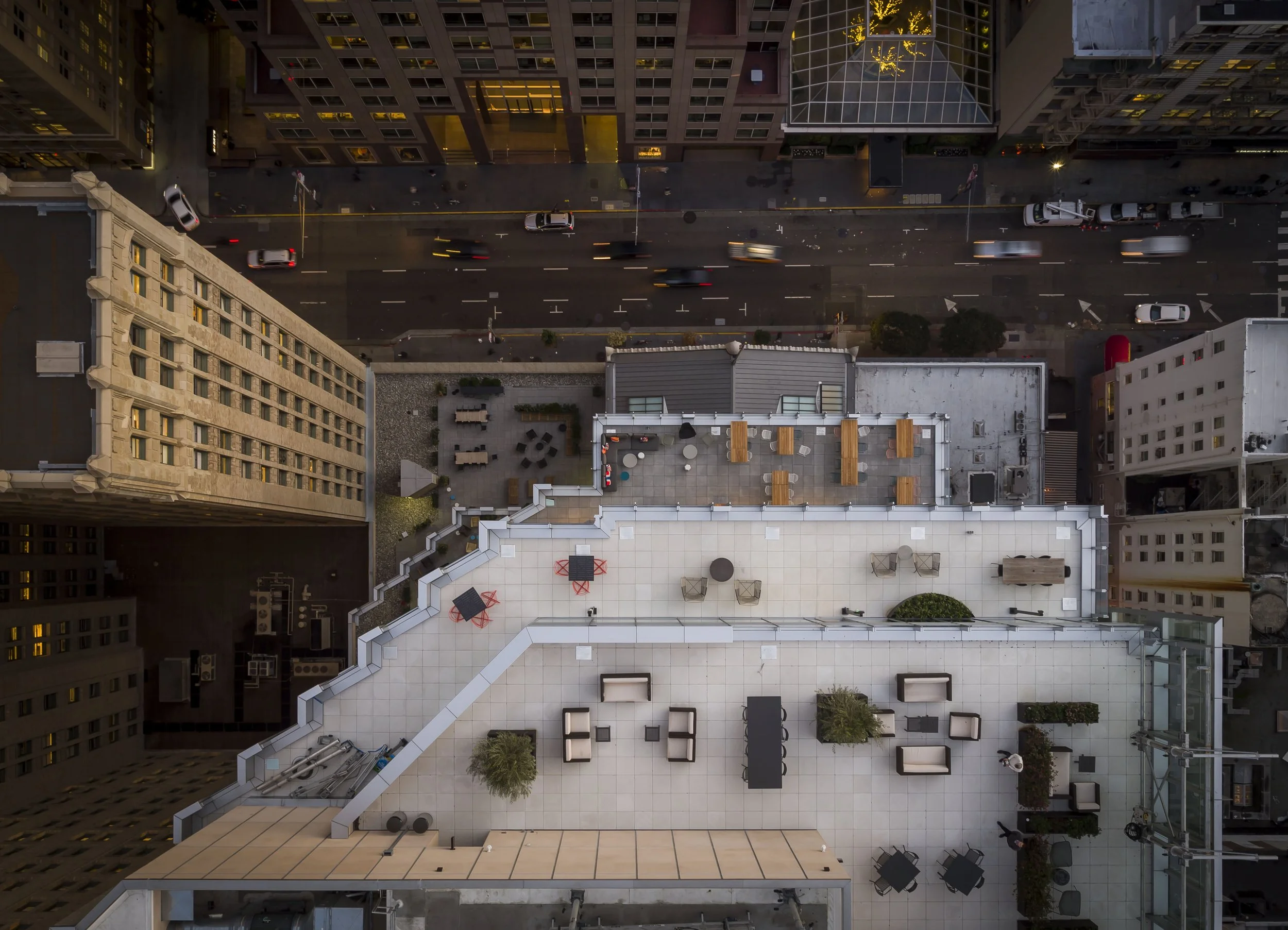 Bird's eye view of a city rooftop patio with outdoor furniture, potted plants, and a view of the city street below with moving cars at night.