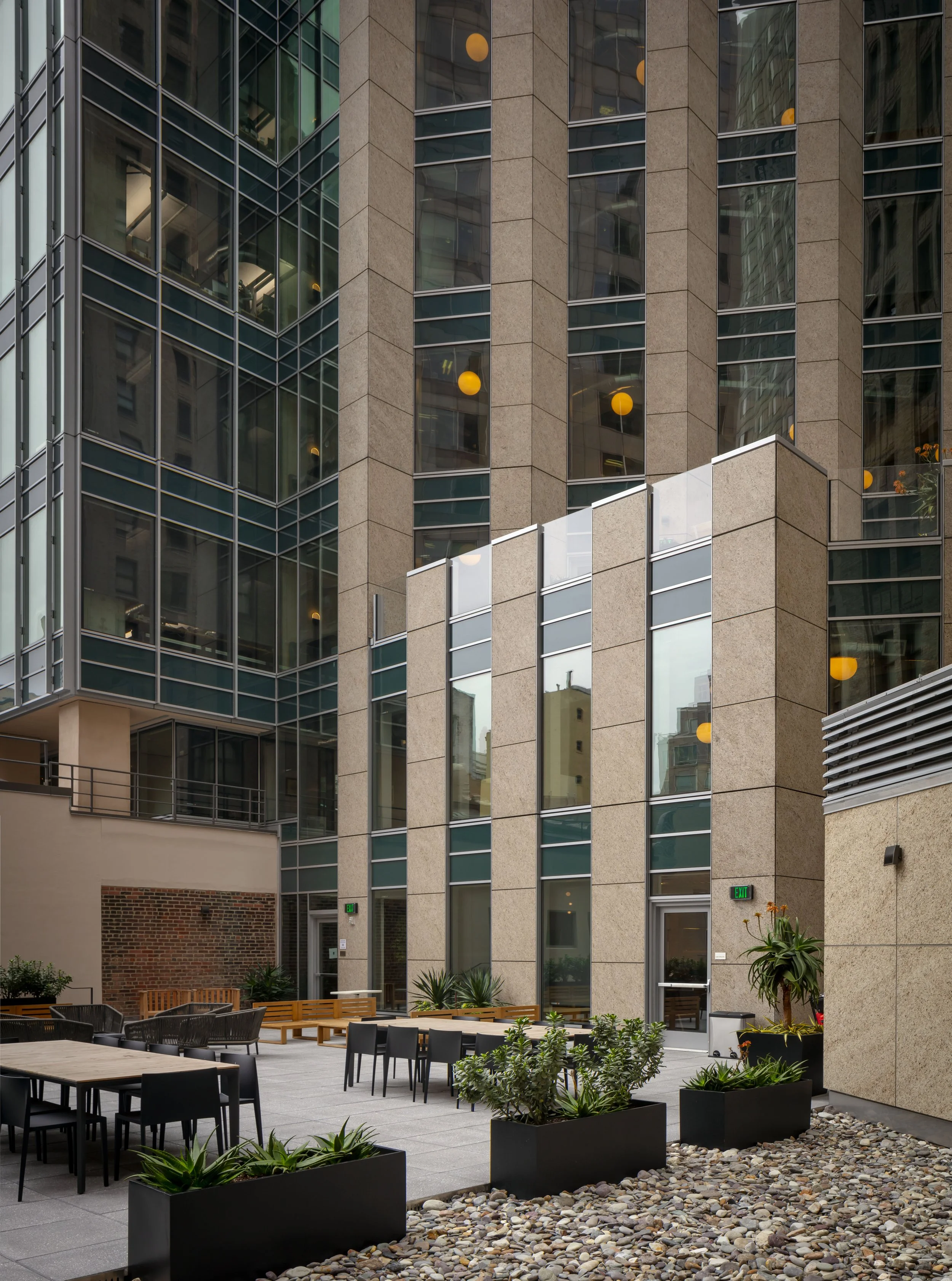 Outdoor seating area with tables, chairs, and planters in front of a modern high-rise building with glass and stone facade.