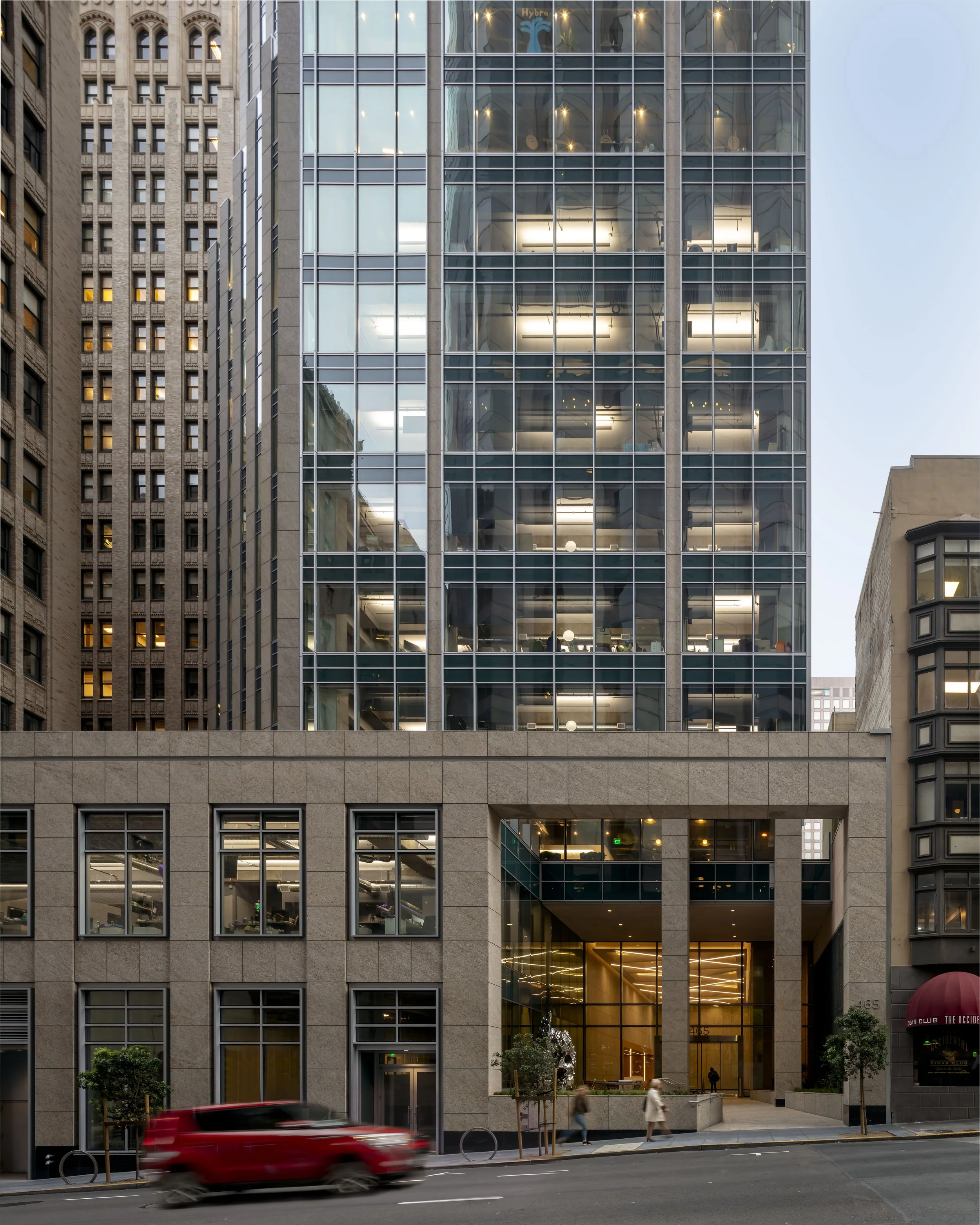 Modern office building with a glass facade reflecting the sky and surrounding structures, with ground-level entrance illuminated with warm lighting, on a city street with a passing red car and pedestrians.