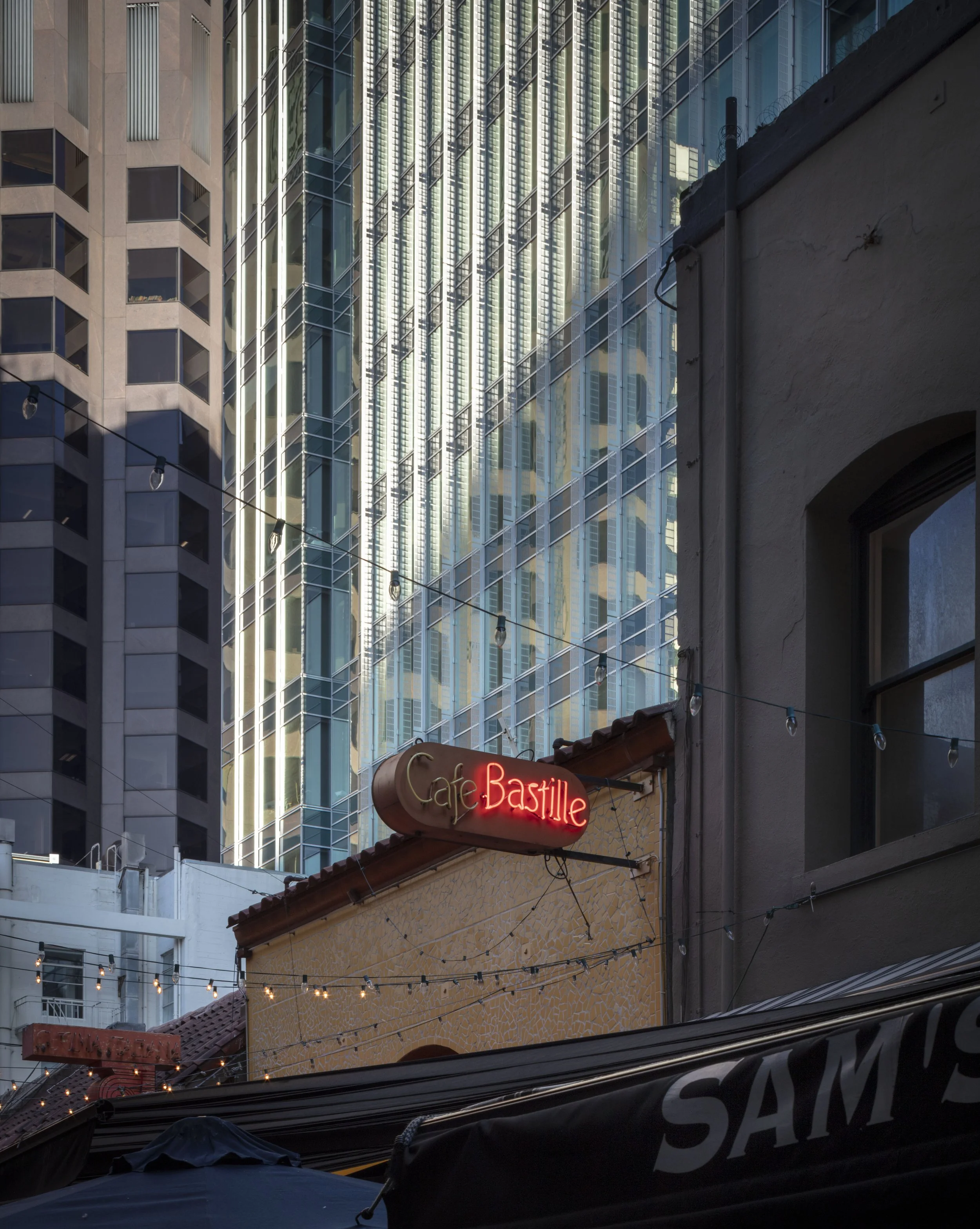 Neon sign that reads 'Cafe Bastille' on a building, with tall modern glass buildings in the background, string lights hanging overhead, and part of a dark awning at the bottom.