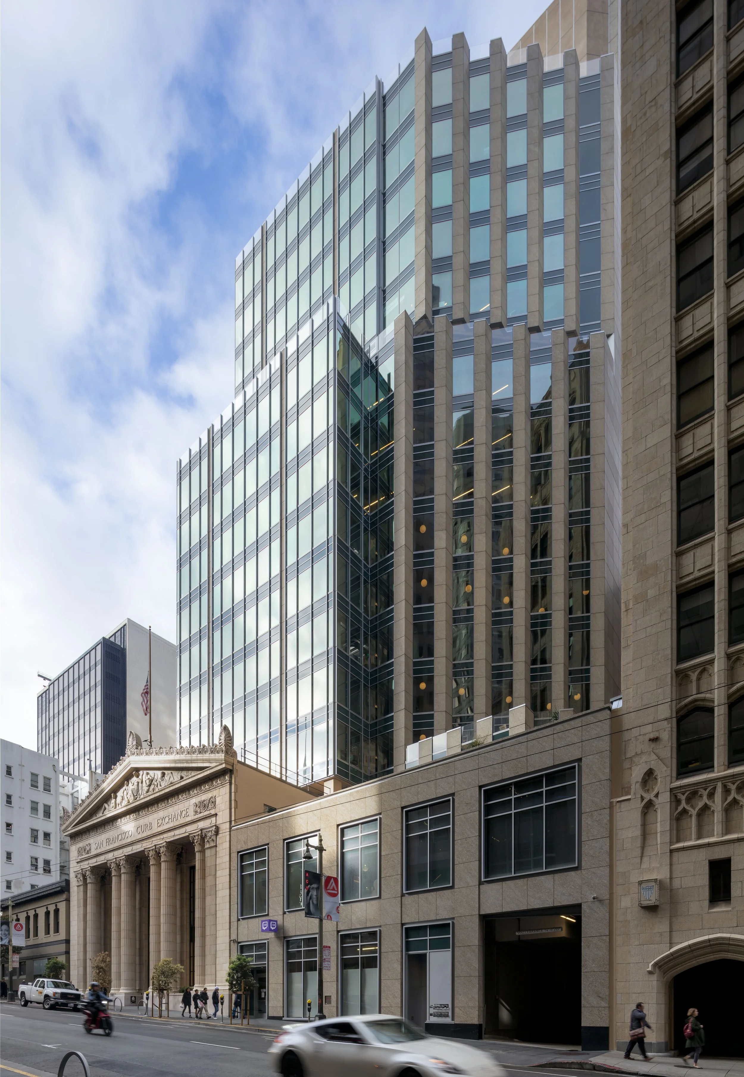 Modern glass skyscraper reflecting the sky and surrounding buildings, with an older neoclassical building in the foreground on a city street.