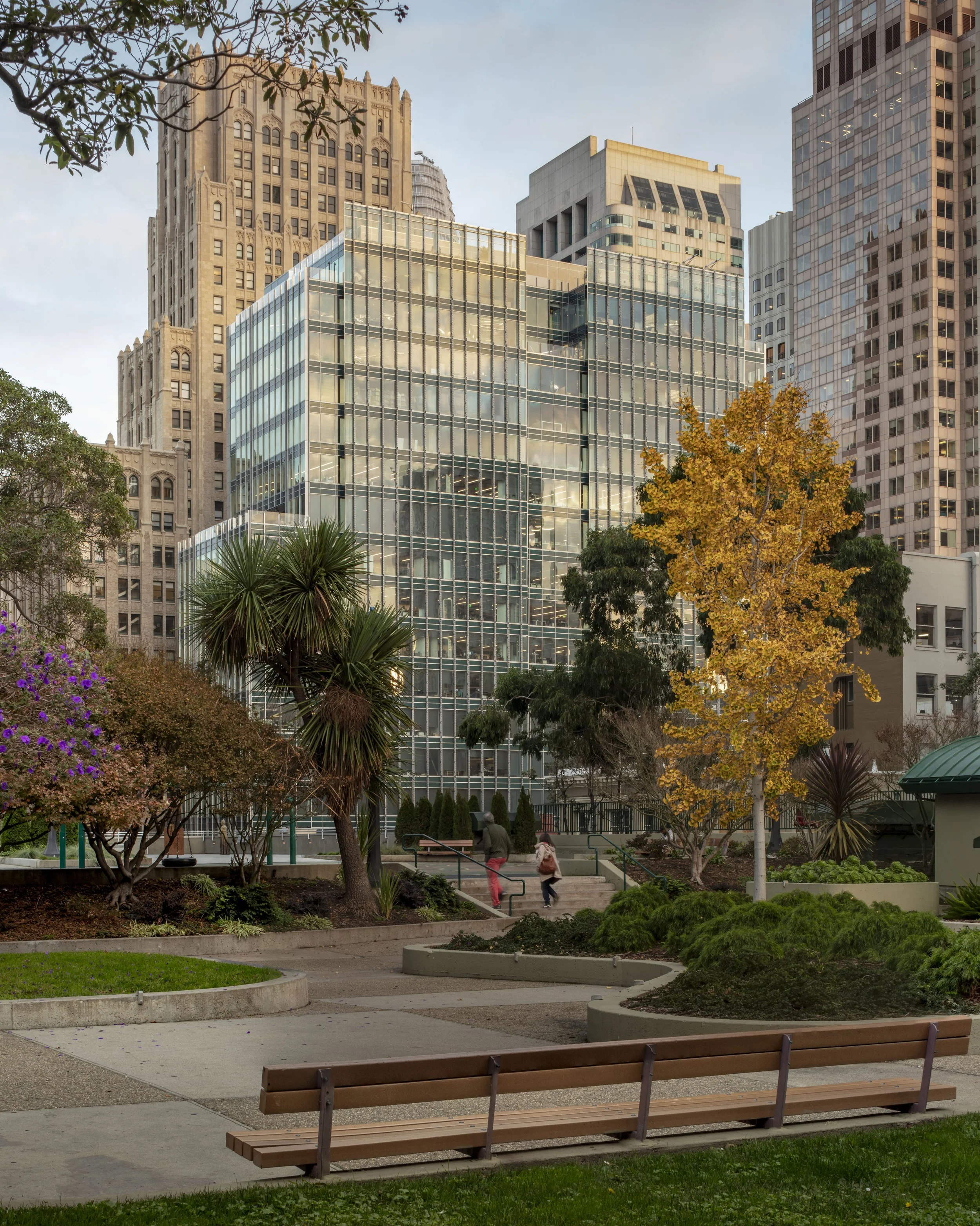 City park with trees, benches, and a modern glass office building in the background during daytime.