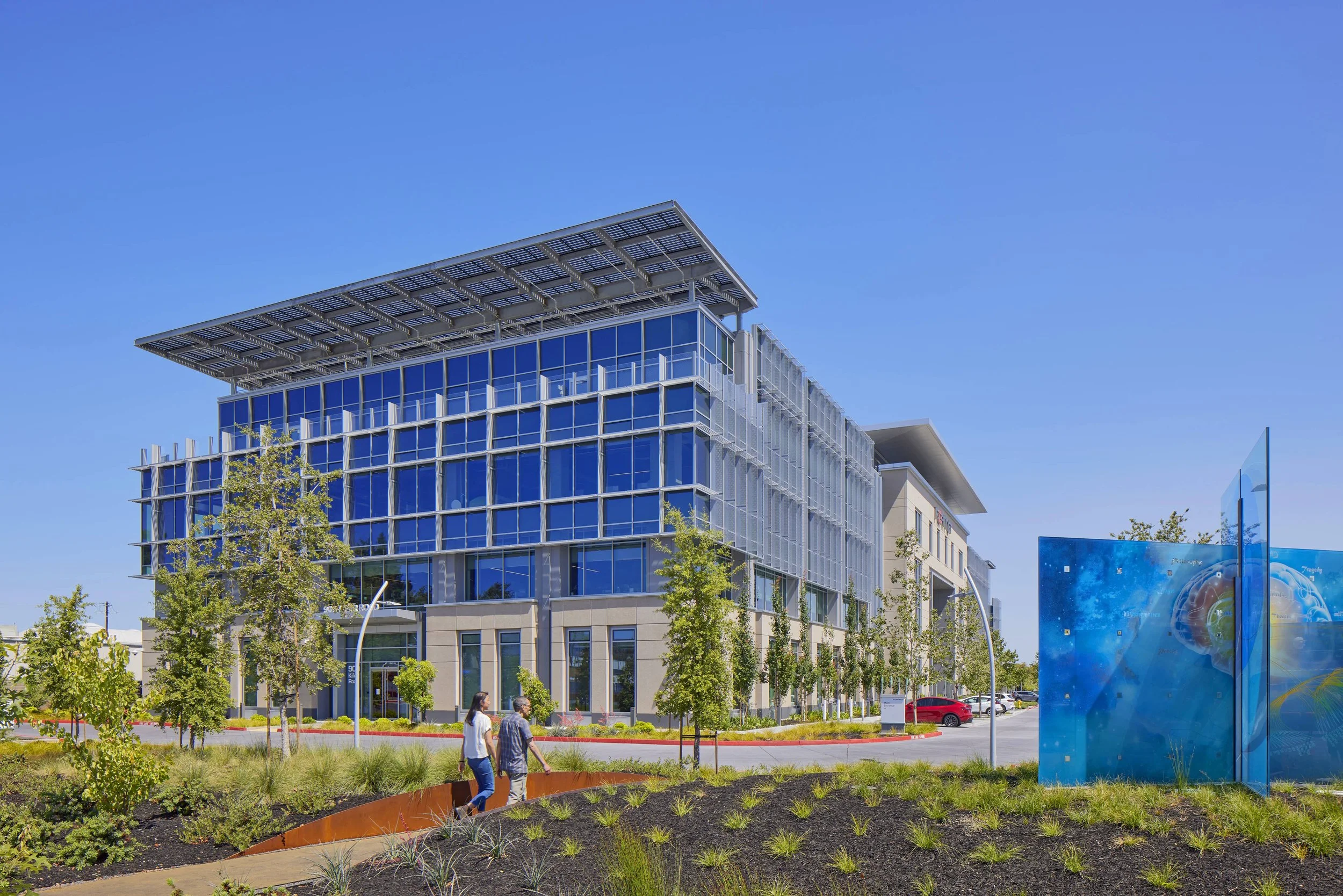 Modern office building with glass windows, surrounded by trees, with a sidewalk and two people walking. Clear blue sky.