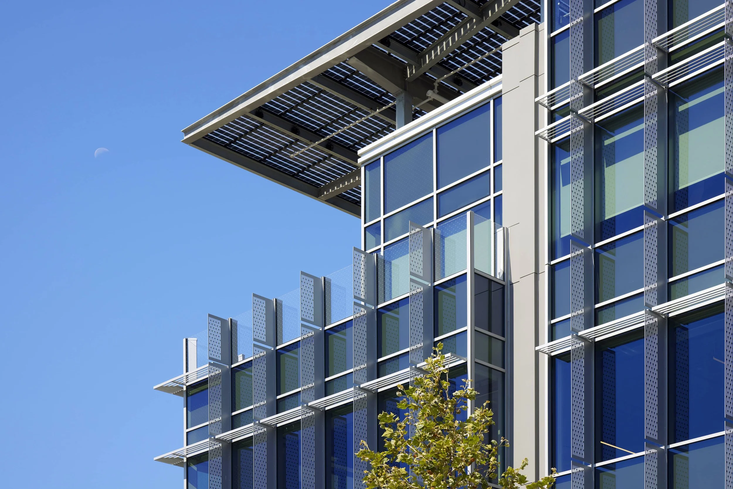Modern glass building with solar panels on a sunny day, with a small moon visible in the blue sky.