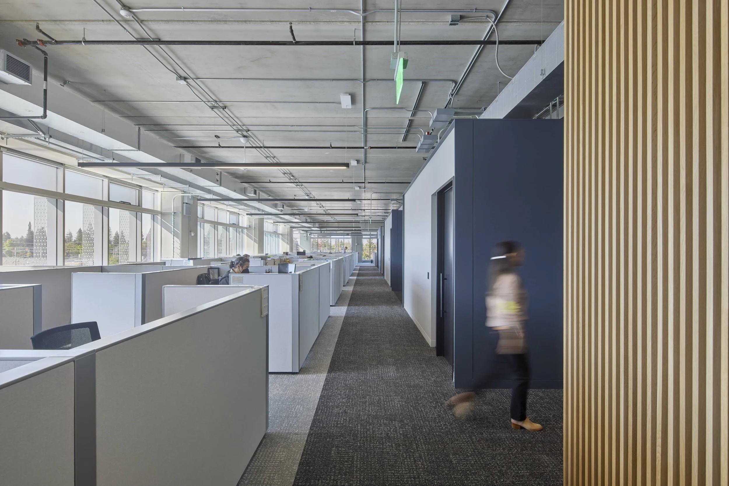Modern office with cubicles along a windowed wall, two employees working at desks, and a woman walking past a wooden slatted wall, with ceiling pipes and ducts visible.