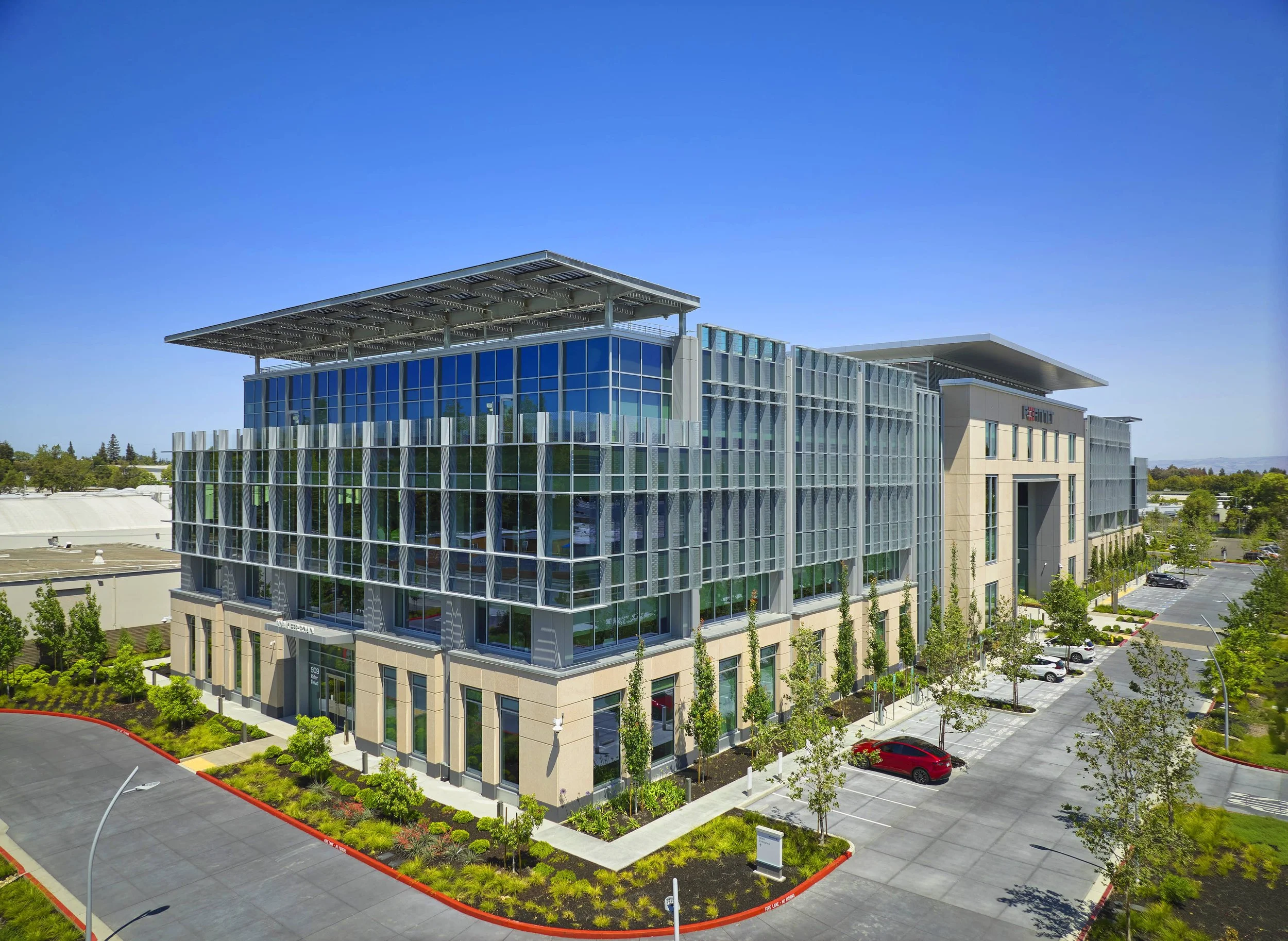 Modern multi-story office building with glass windows, surrounded by parking lot and landscaped greenery, under clear blue sky.
