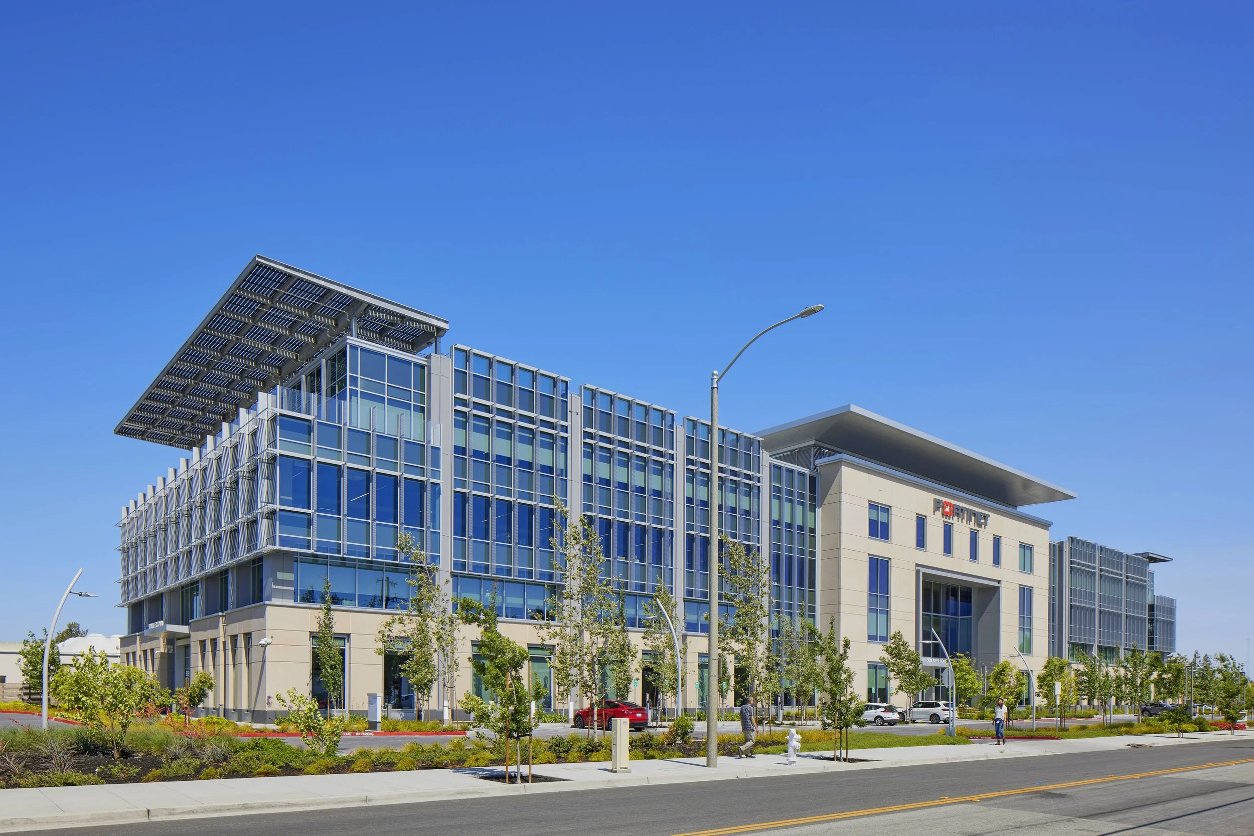 Modern office building with glass and concrete exterior, trees, and cars parked in front, under a clear blue sky.
