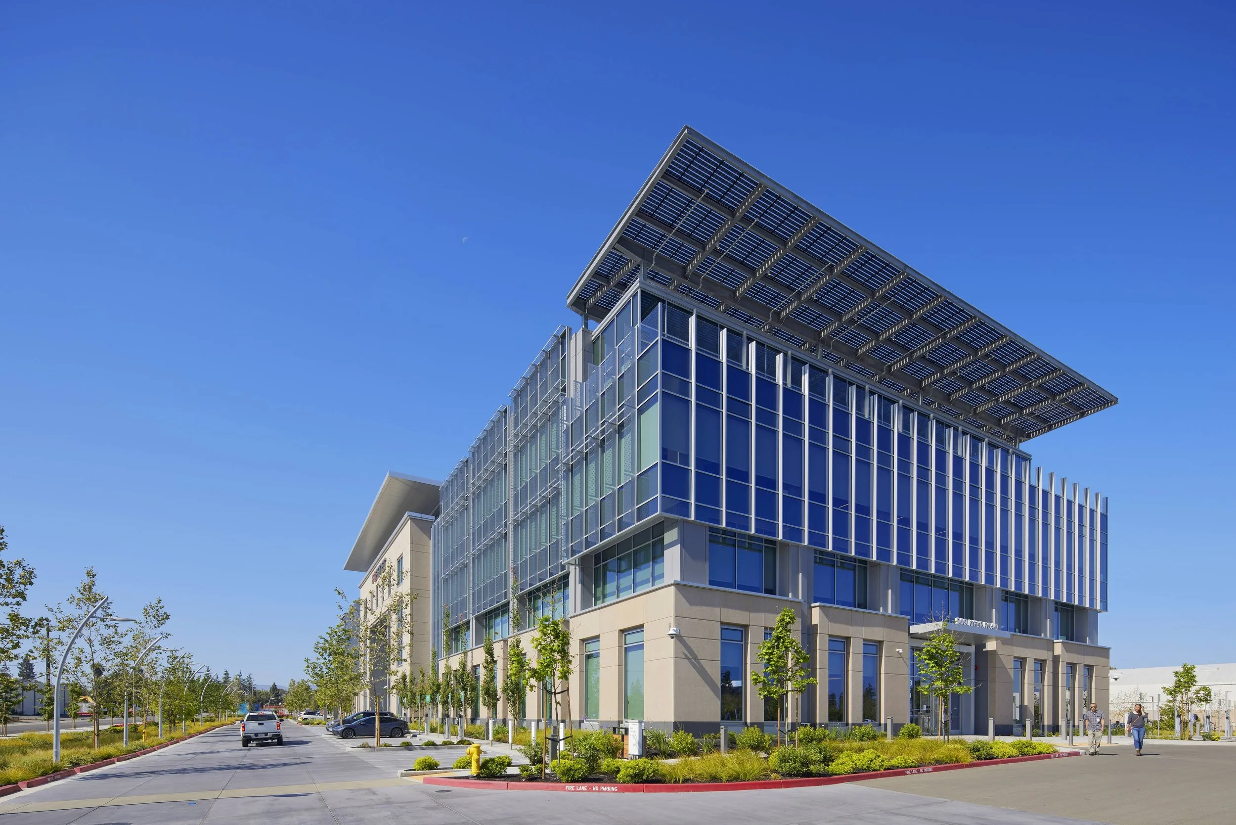 Modern multi-story office building with glass facade and solar panels on the roof, surrounded by a parking lot and small trees, under a clear blue sky.