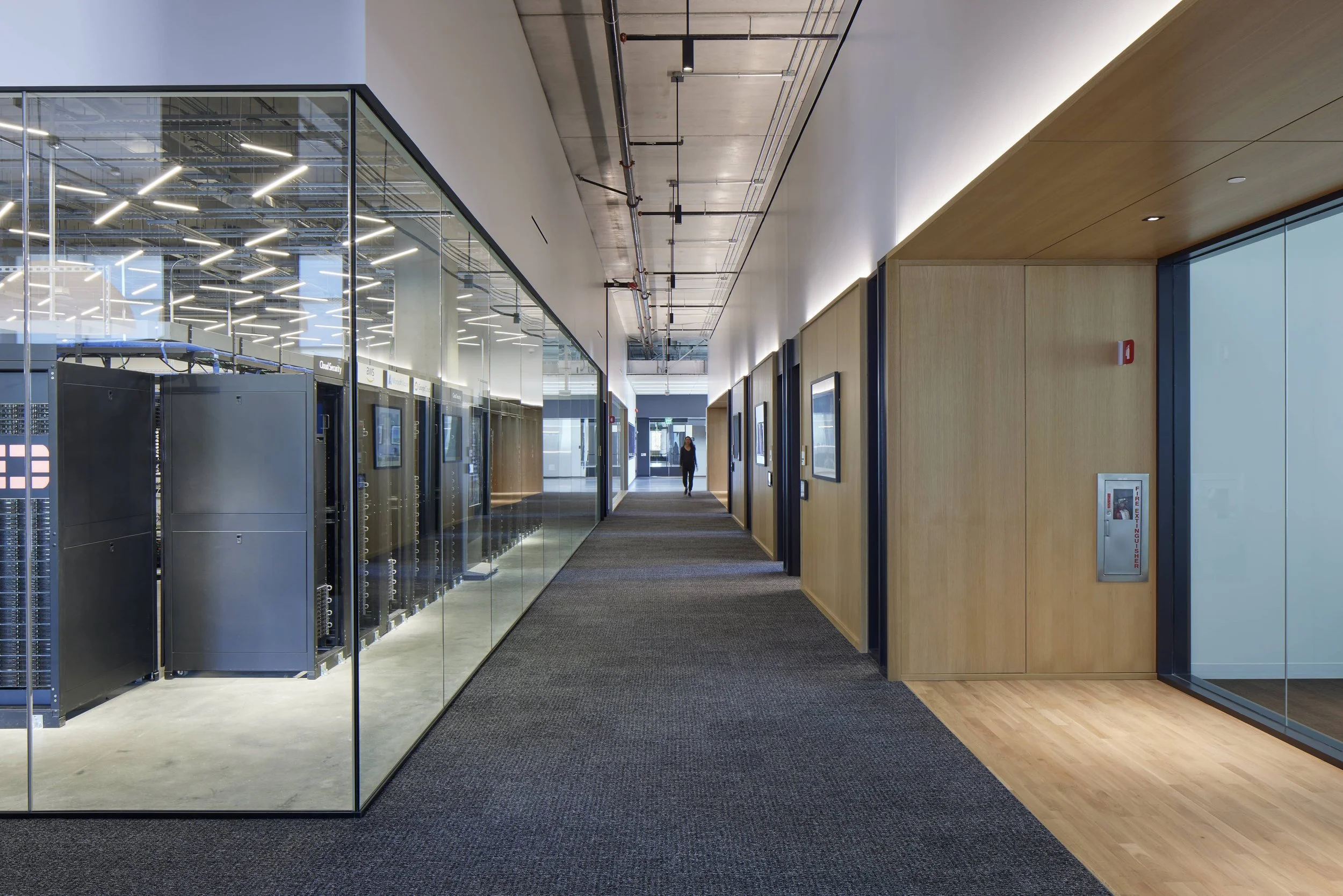 Modern office corridor with glass-walled server room on the left and wooden-paneled walls on the right, with a woman walking in the distance.