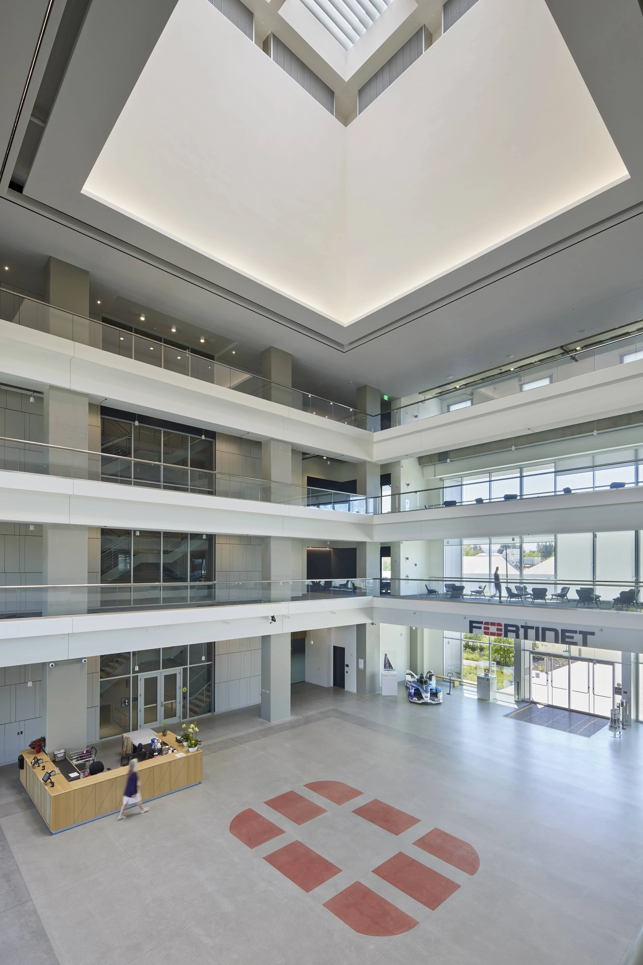 Interior view of a modern office building lobby with multiple floors, large glass windows, a reception desk, and a spacious open area with a Fortinet sign near the entrance.