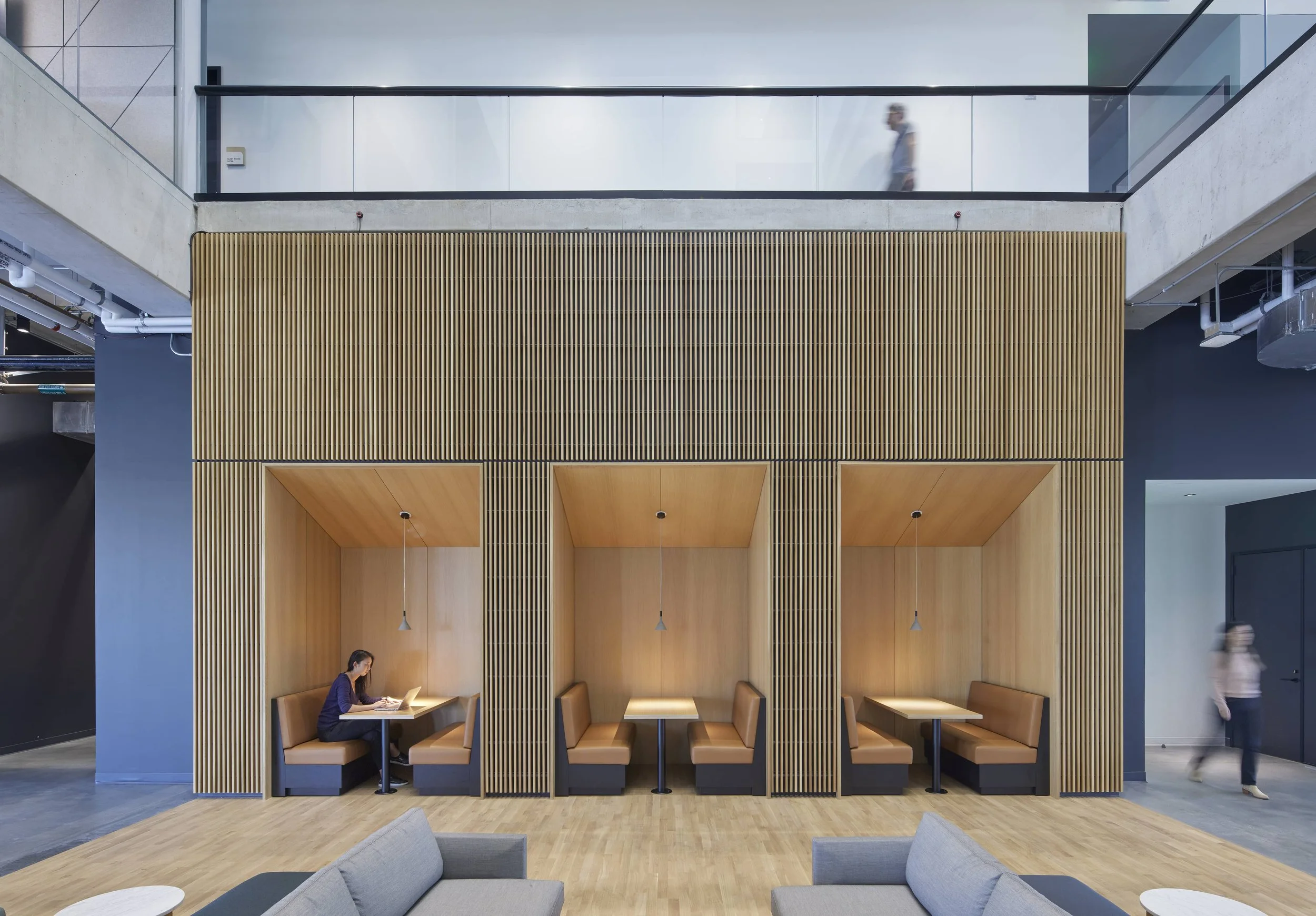Modern interior of a building with three private booth seating areas with wooden paneling, a woman working on a laptop in the left booth, and two blurred people walking in the background.