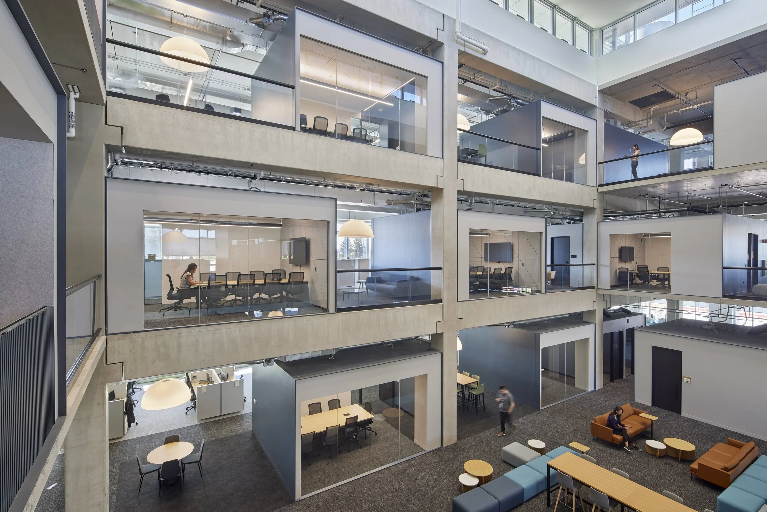 Modern office building with open and closed meeting rooms, glass walls, and multiple floors. Visible people working and walking, with office furniture and seating areas.