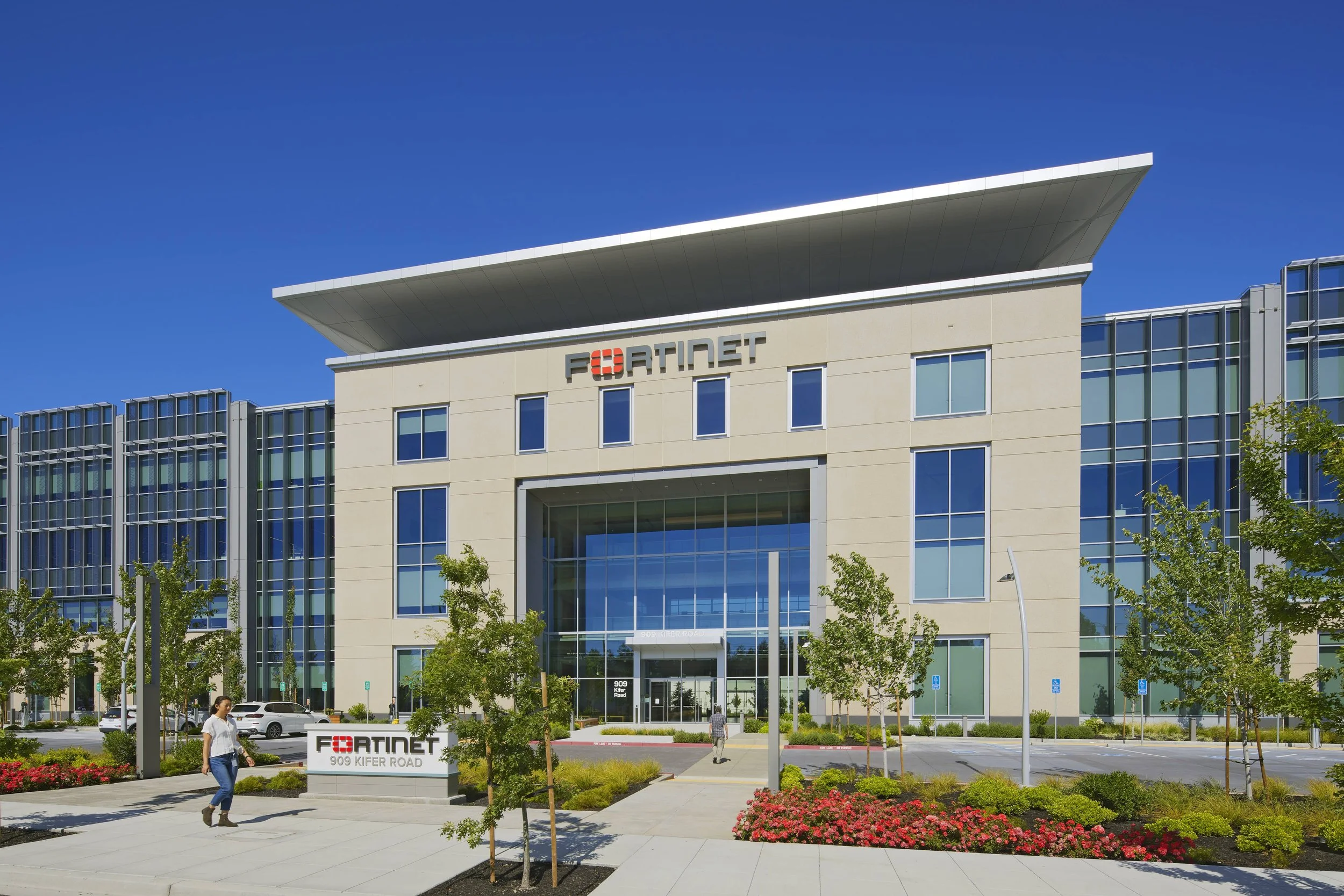 Front view of the Fortinet building with a clear blue sky, modern glass windows, landscaping, and people walking.
