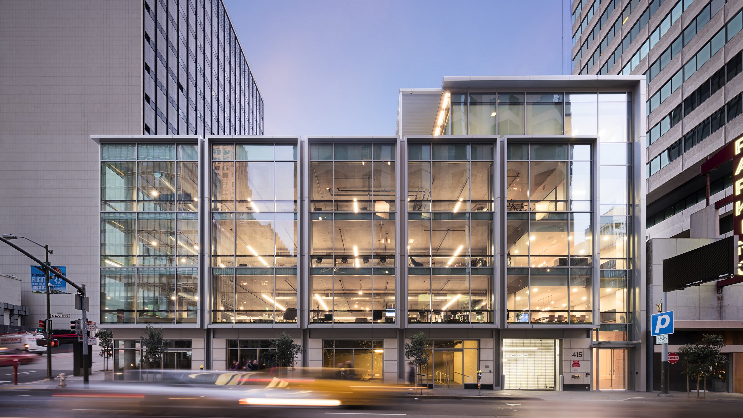 Modern building with large glass windows on a city street at dusk, with visible interior lighting and people inside.