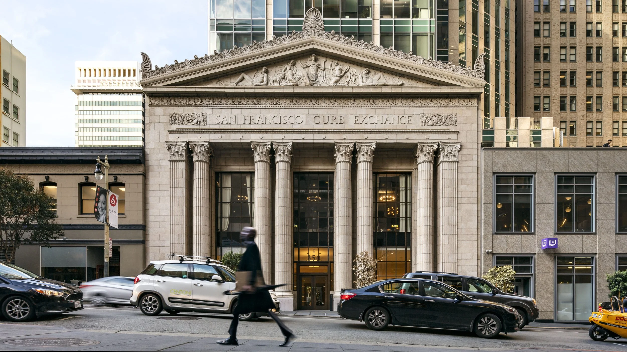 Facade of the San Francisco Curb Exchange building with classical columns and intricate architectural details, situated in an urban street scene with cars, pedestrians, and modern high-rise buildings in the background.