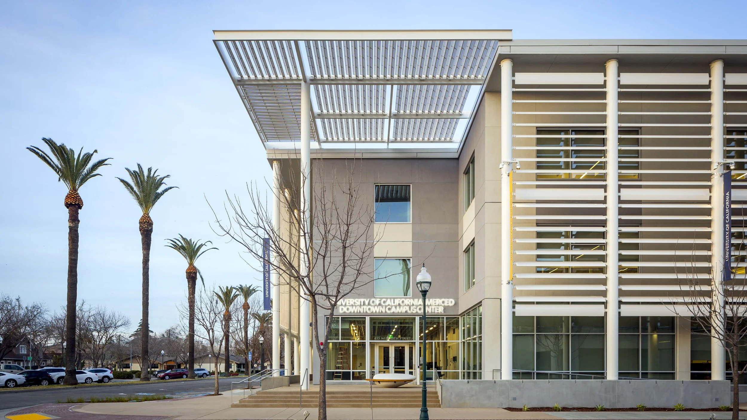 Exterior of the University of California Merced Downtown Campus Center with palm trees, leafless trees, a lamp post, and a building with large glass windows.