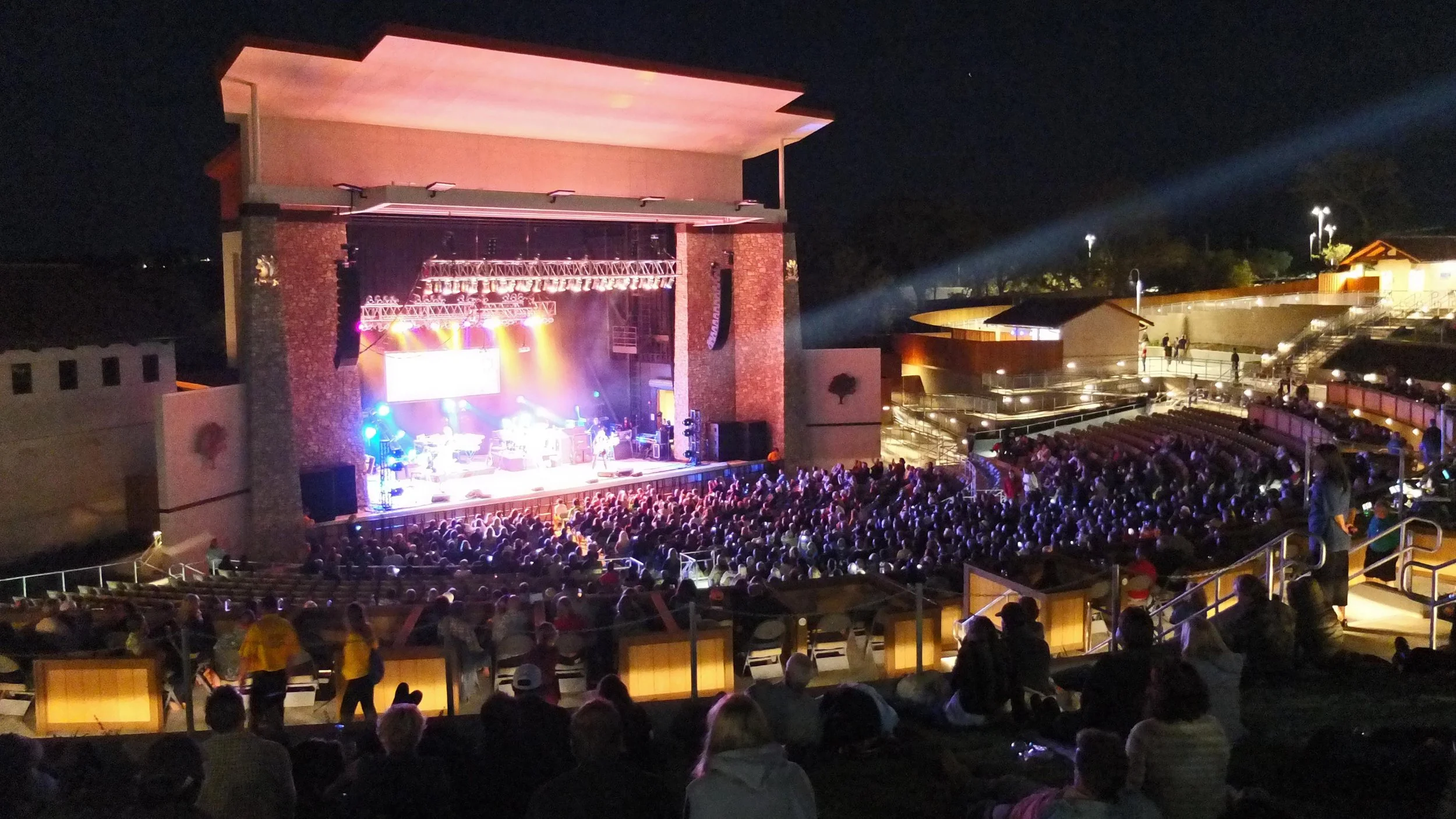 An outdoor concert at night with a large stage illuminated with colorful lights, a band performing, and a packed audience seated in tiered seating and standing areas.