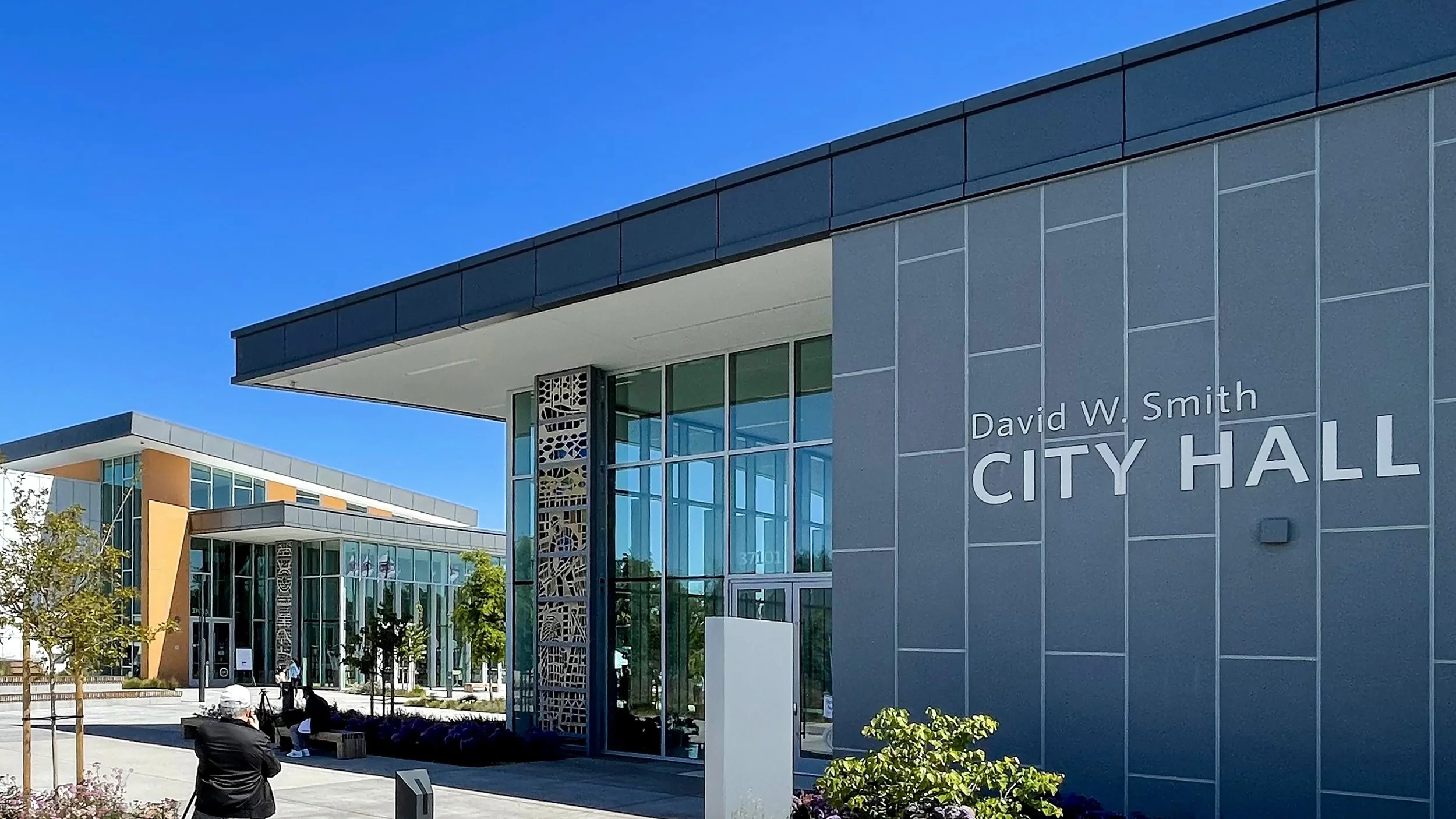 Modern city hall building with glass windows, trees, and people outside on a sunny day.