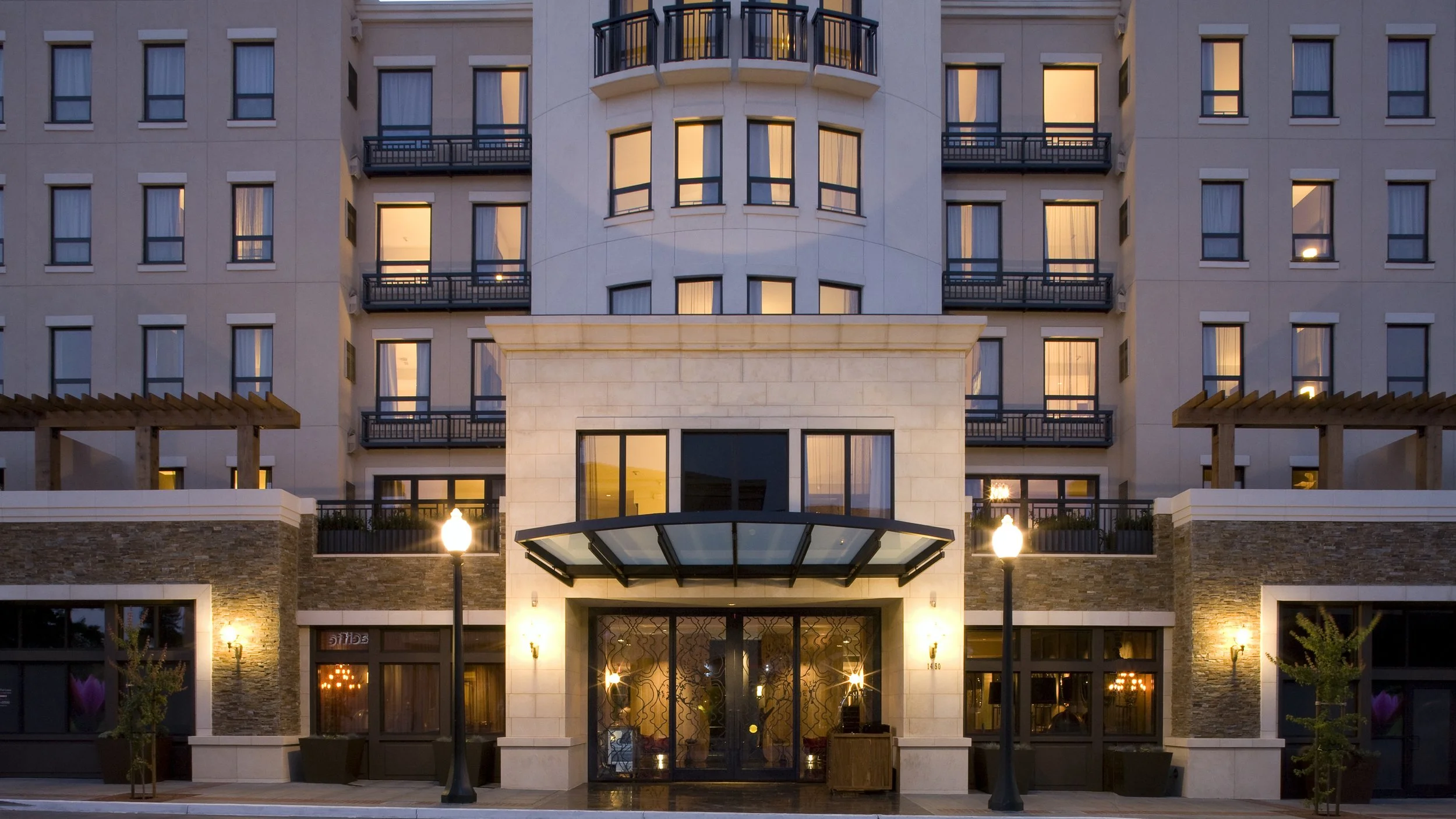 Front view of a modern multi-story hotel building with illuminated windows, entrance with glass doors, and street lamps at dusk.