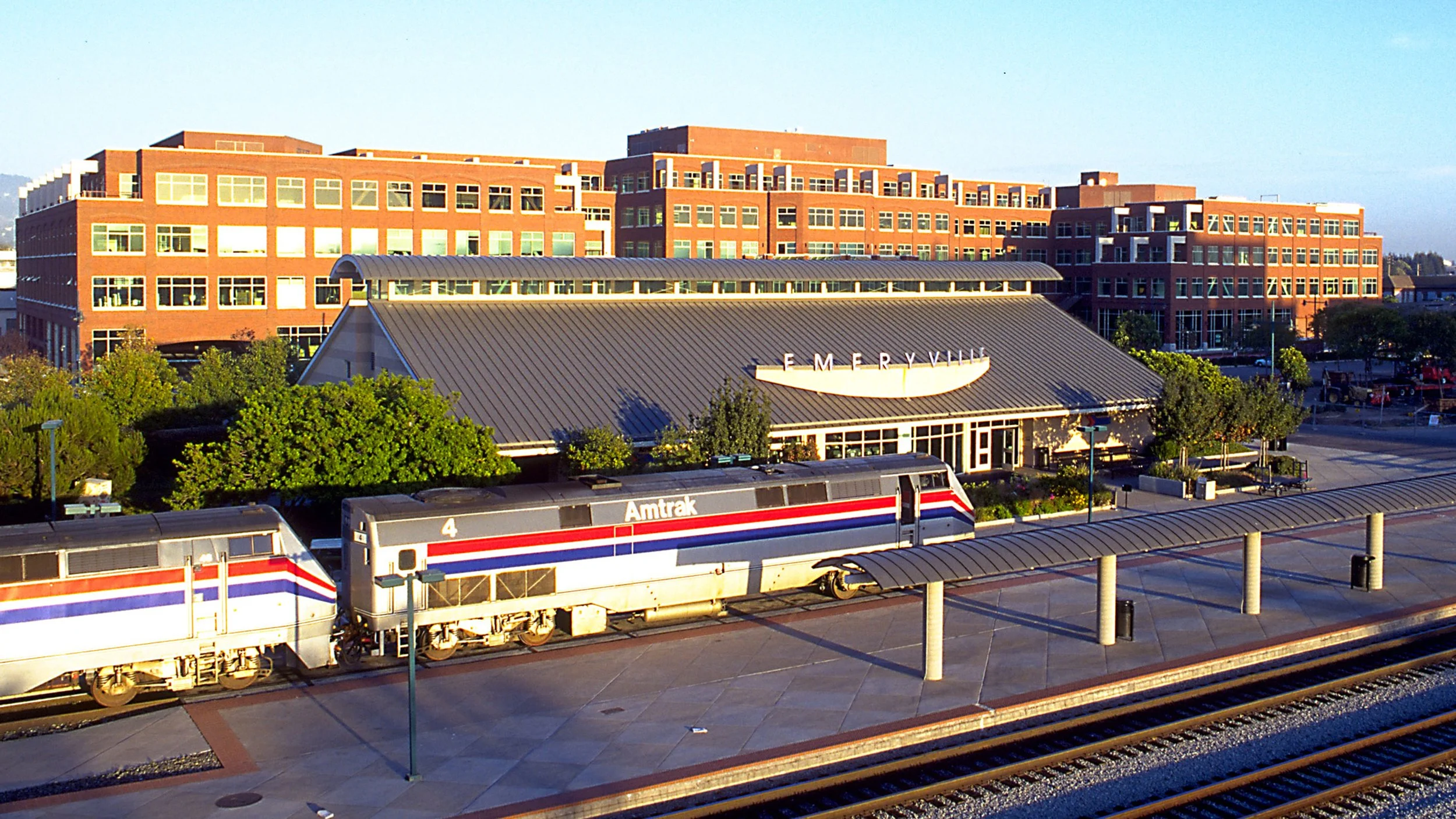 Amtrak train at a station with a mall in the background, in the late afternoon sunlight.