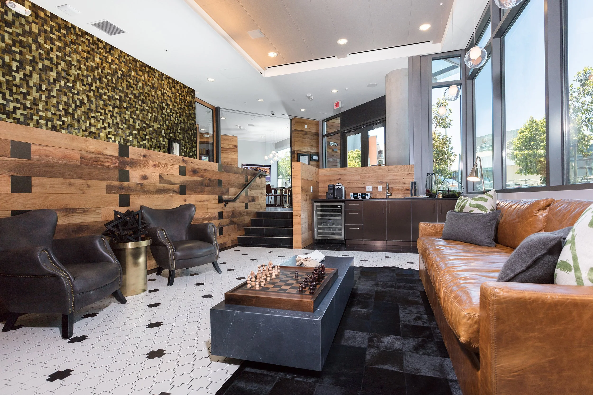Modern lobby area with leather sofa, armchairs, chessboard on a black marble coffee table, large windows, wooden accents, and a kitchenette in the background.