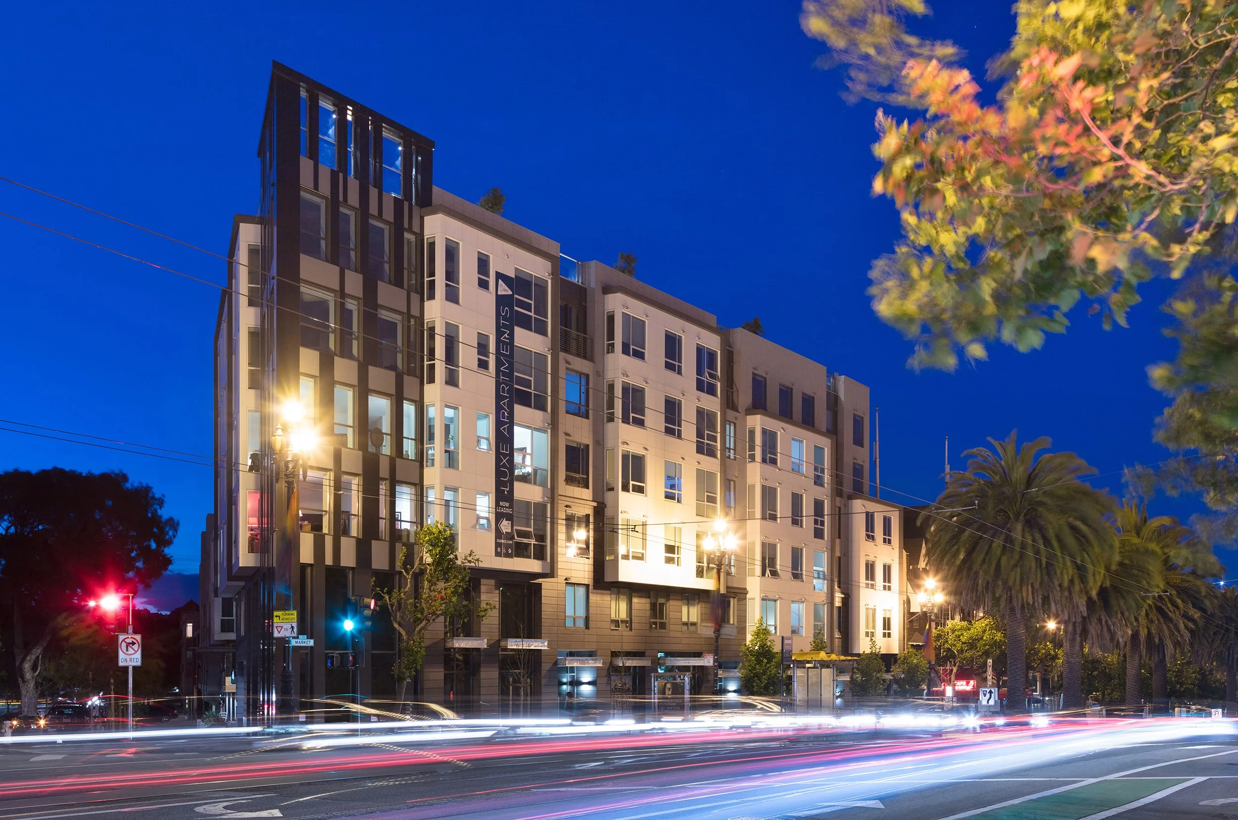 Nighttime city street with a modern multi-story apartment building lit up and cars passing, leaving light trails, with trees and streetlights in the foreground.