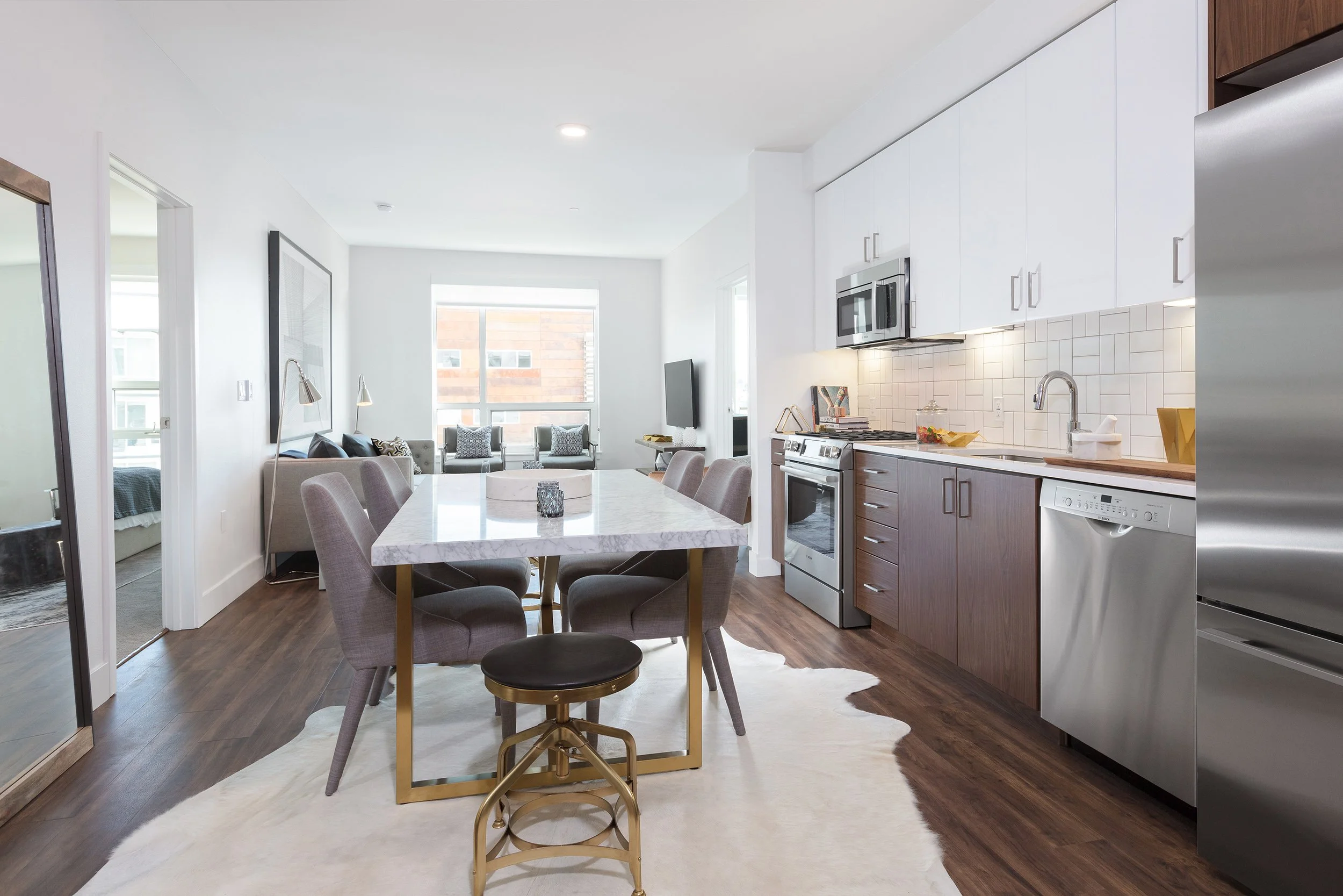 Open-concept kitchen and living room with white cabinets, stainless steel appliances, a marble dining table, and modern upholstered chairs, with a cowhide rug and large windows.